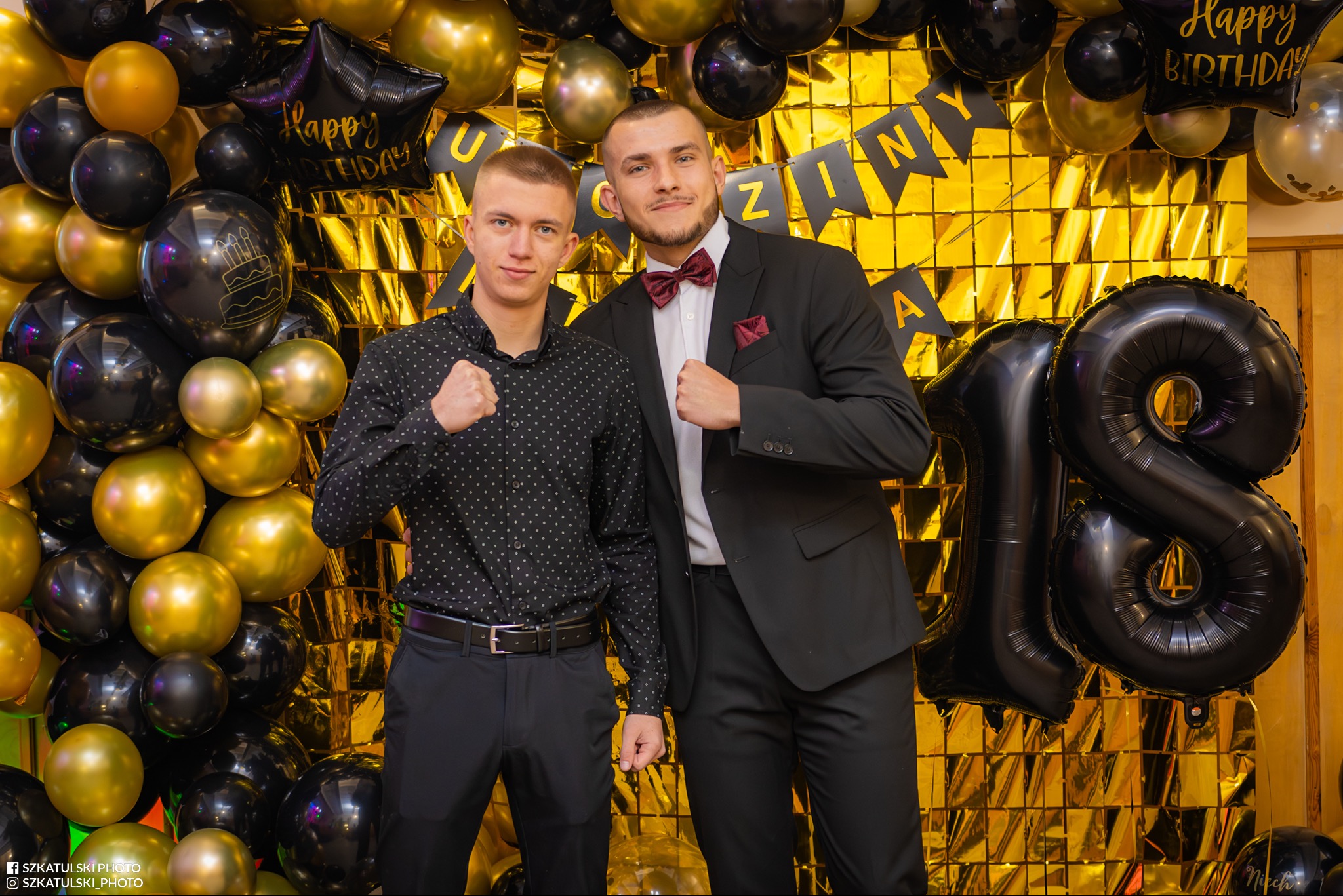 Dwóch eleganckich mężczyzn pozuje na tle dekoracji urodzinowych z balonami i złotą ścianką. Jeden w garniturze z muchą, drugi w koszuli. Uśmiechy i radosna atmosfera.