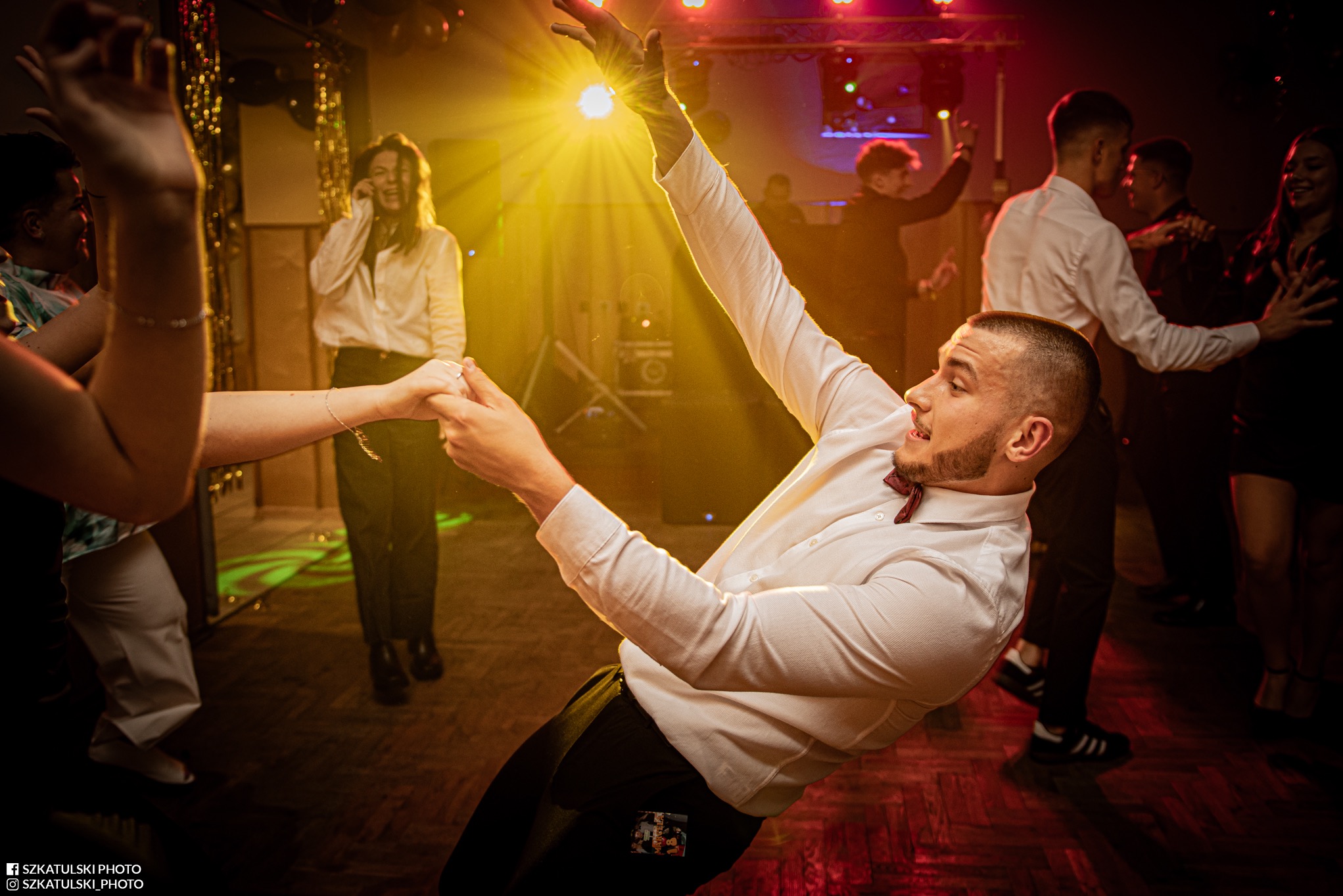 Roztańczony mężczyzna w białej koszuli i muszce, trzymający kogoś za rękę na parkiecie. W tle bawiący się goście i światła dyskotekowe.