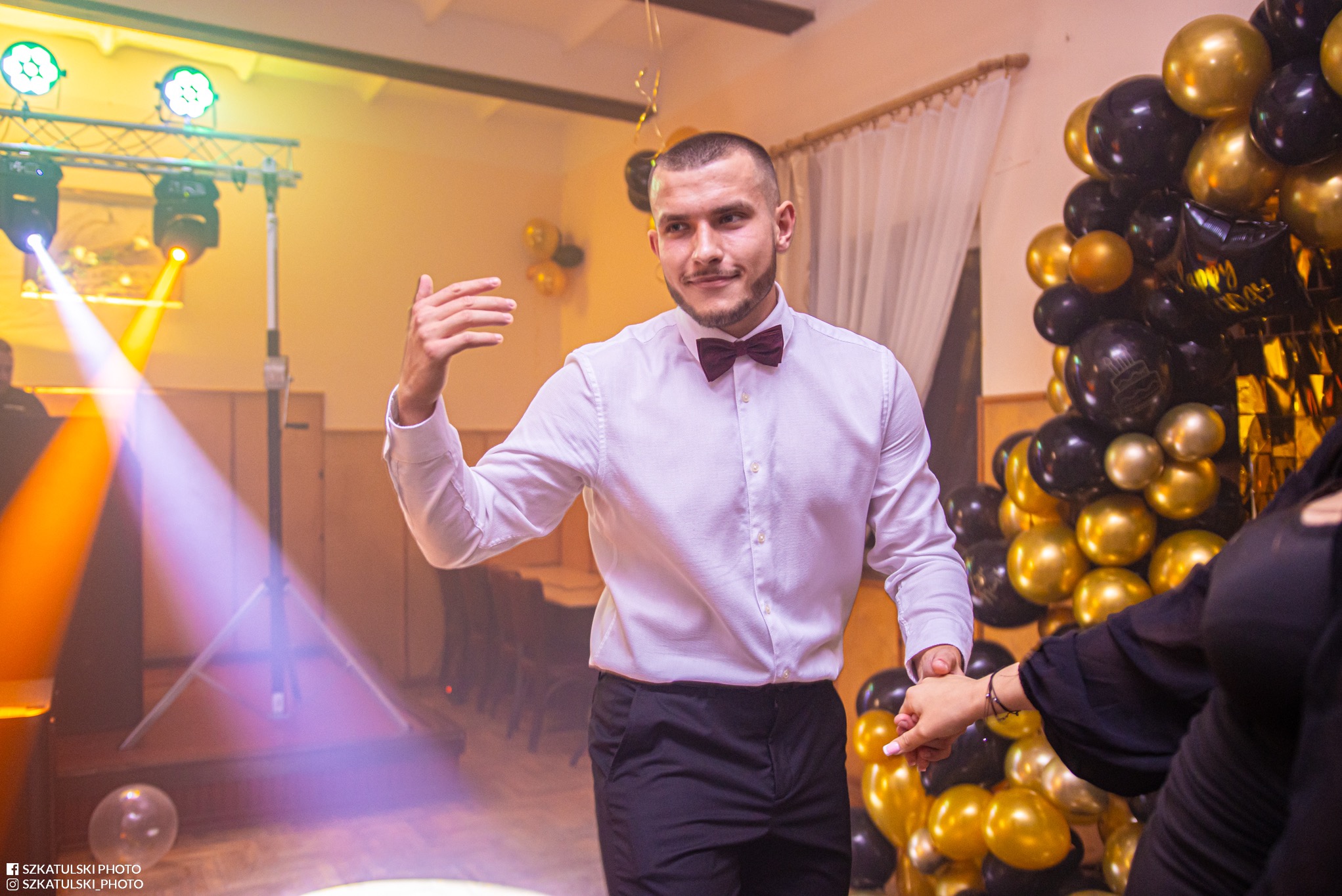 Mężczyzna w muszce gestykuluje na tle dekoracji z czarno-złotych balonów i oświetlenia scenicznego podczas imprezy. Widoczna dłoń kobiety.