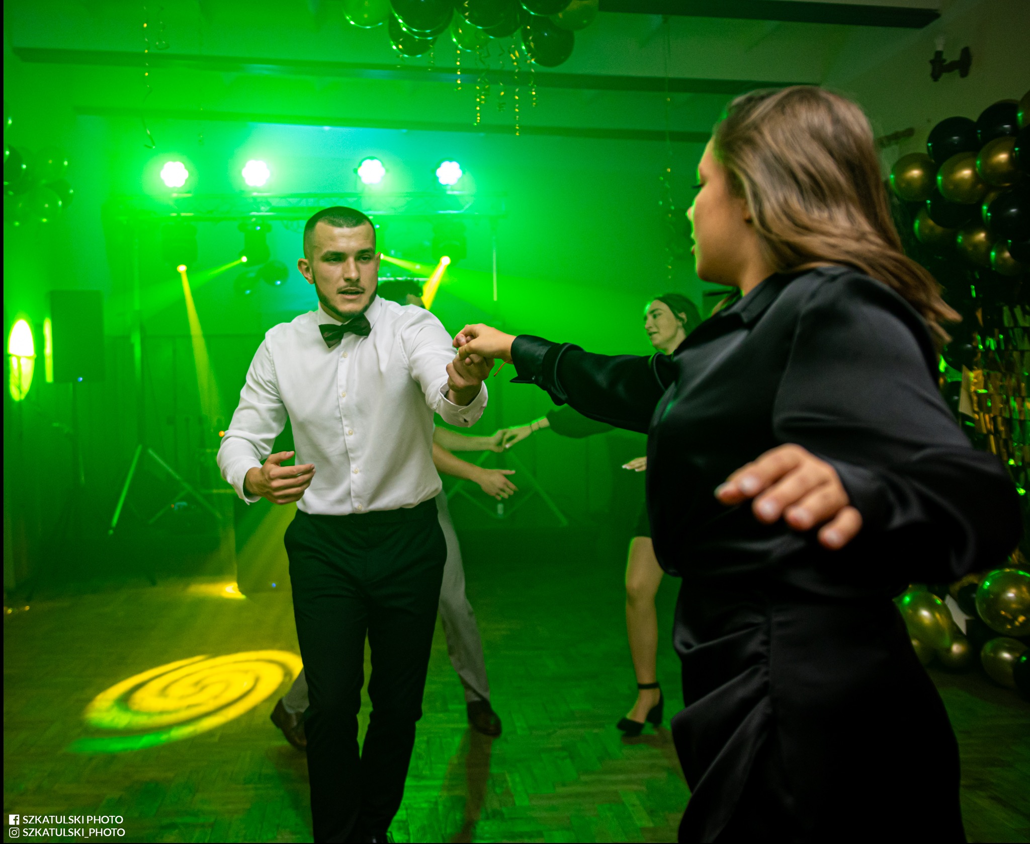 Para trzymająca się za ręce na parkiecie podczas imprezy. W tle widoczne dekoracje z balonów i zielone światła. Mężczyzna w białej koszuli i muszce, kobieta w czarnej sukience.