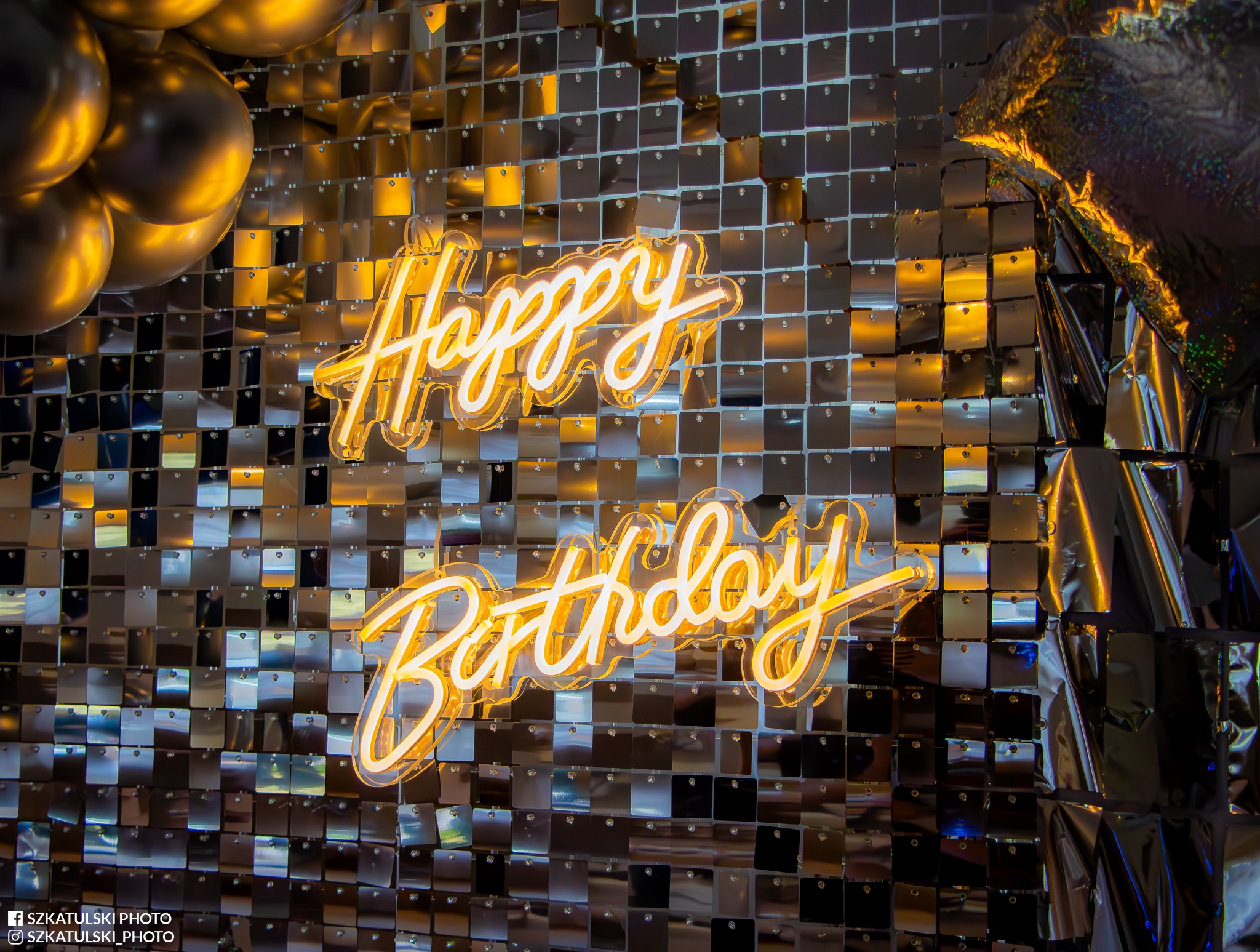 Neonowy napis 'Happy Birthday' na błyszczącym tle z cekinów i balonów. Dekoracja urodzinowa w ciepłych, złotych barwach. Ujęcie z bliska, podkreślające detale.