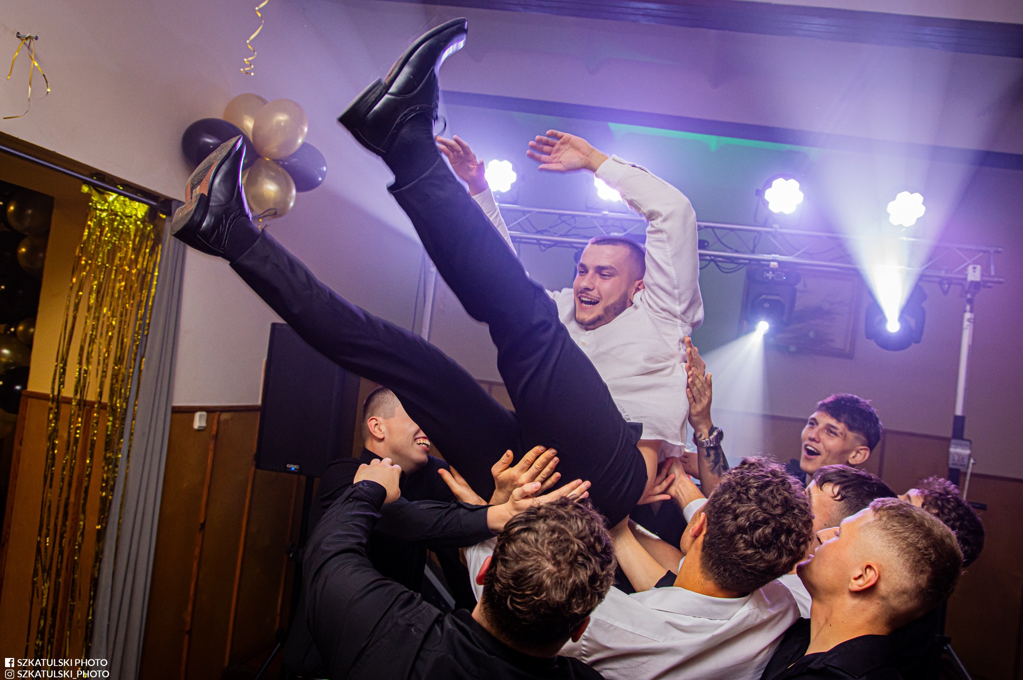 Radosny mężczyzna w białej koszuli podrzucany przez grupę osób na imprezie. W tle balony i oświetlenie sceniczne. Dynamiczne ujęcie z perspektywy żabiej.