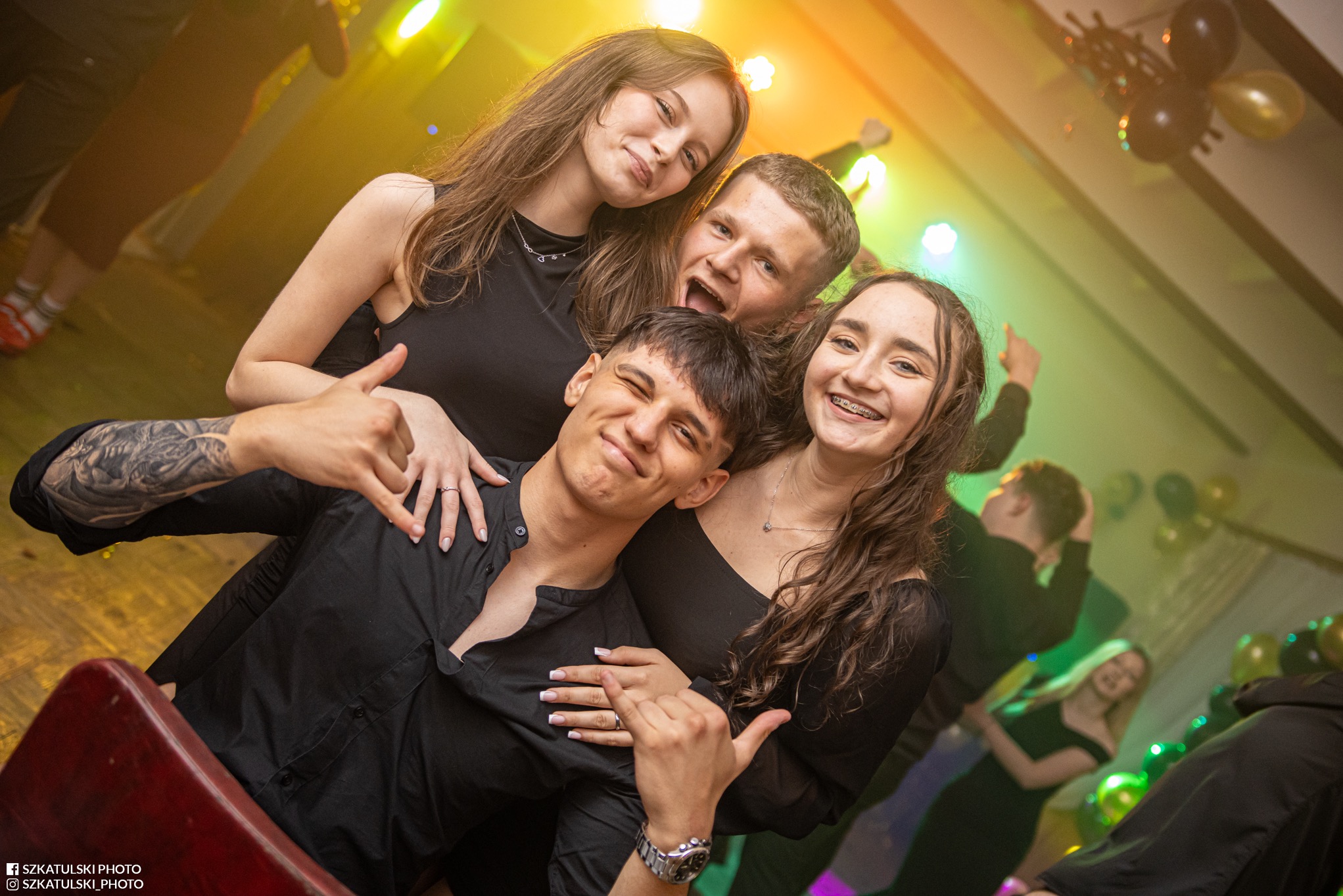 Radosna grupa czterech młodych osób w czarnych strojach pozuje do zdjęcia na tle dekoracji balonowych i świateł na imprezie. Uśmiechy, bliskość i pozytywna energia.