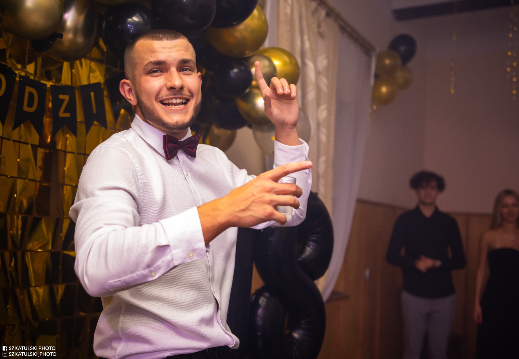 Uśmiechnięty mężczyzna w muszce, wznosi toast na tle dekoracji z czarno-złotych balonów i błyszczącej kurtyny. W tle widoczne dwie osoby.