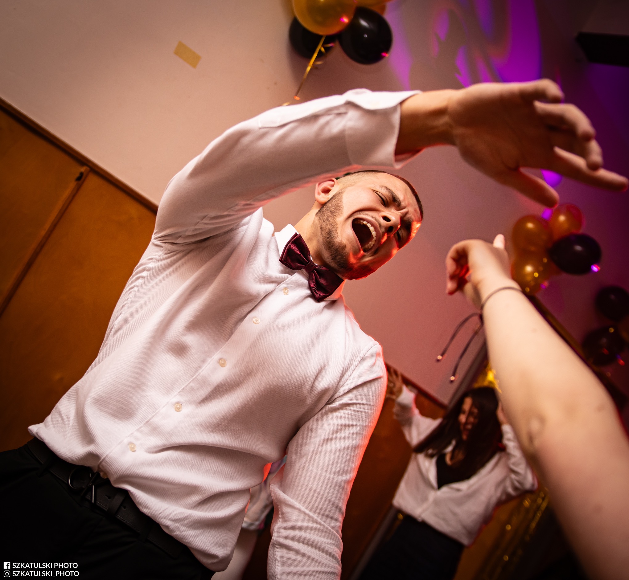 Rozbawiony mężczyzna w białej koszuli i bordowej muszce, z otwartymi ustami, tańczy na imprezie. W tle balony i inne osoby. Dynamiczne ujęcie z dołu.