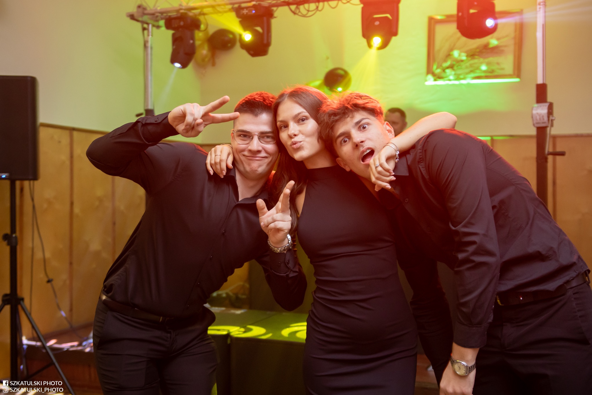 Radosne ujęcie trzech osób w Sulejowie, pozujących do zdjęcia z gestami. Oświetlenie sceniczne w tle, ciemne stroje. Fotografia eventowa.