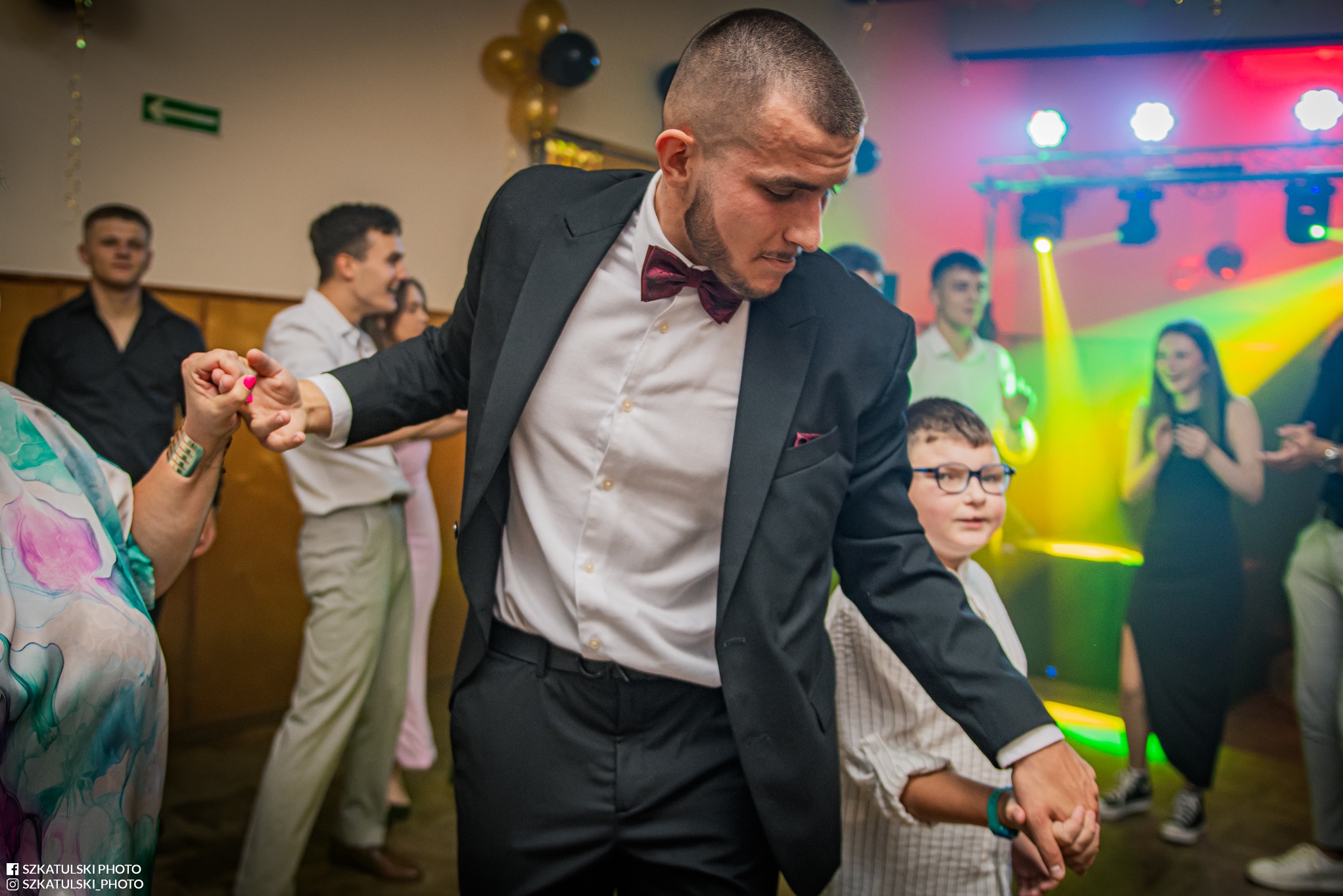 Mężczyzna w garniturze tańczy, trzymając za rękę chłopca w okularach na tle rozświetlonej sali balowej z innymi osobami. Ujęcie z dołu, dynamiczne światło.