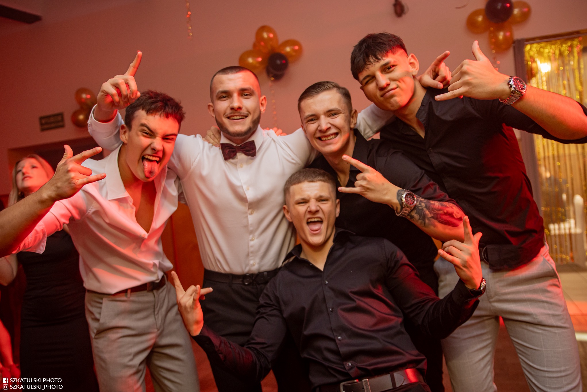 Rozbawiona grupa młodych mężczyzn w eleganckich strojach pozuje do zdjęcia na tle dekoracji imprezowej, gestykulując i uśmiechając się.