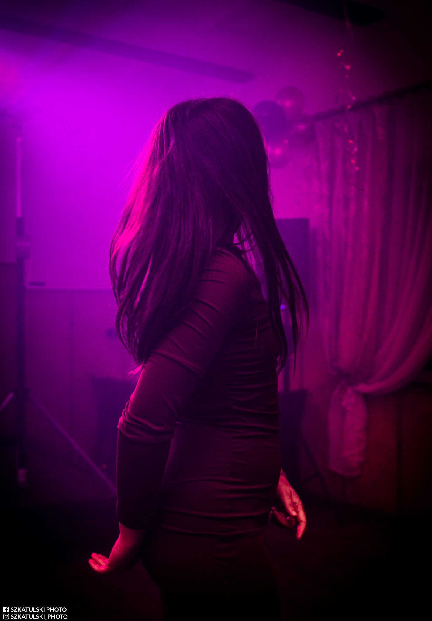 Kobieta w ciemnej sukience, odwrócona tyłem, w fioletowym oświetleniu. W tle widoczne balony i zasłony. Ujęcie z dołu, dynamiczne, z efektem ruchu włosów.
