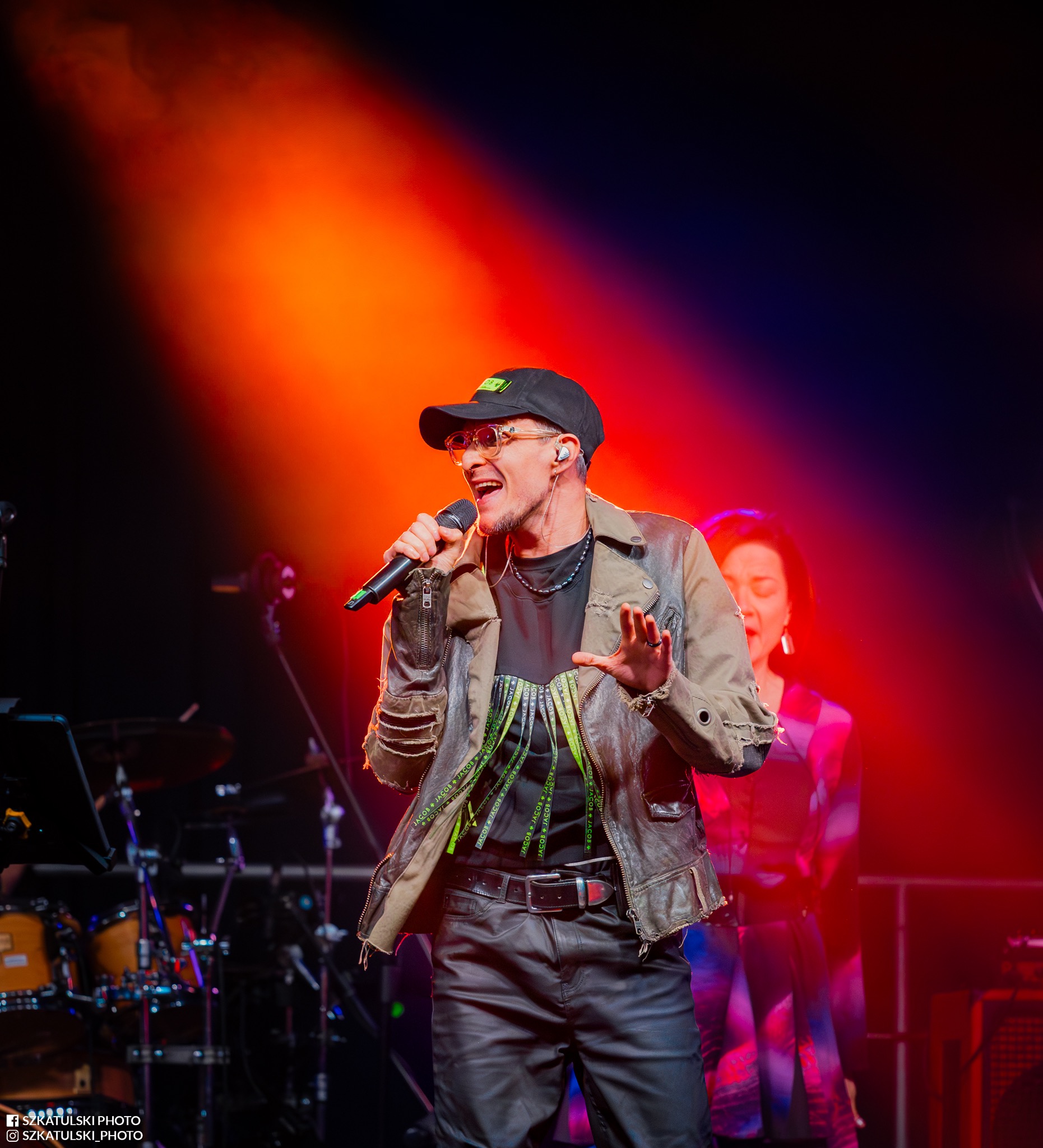 Wokalista w skórzanej kurtce, czapce i okularach śpiewa do mikrofonu na scenie oświetlonej czerwonym światłem. W tle widoczna perkusja i postać kobiety.