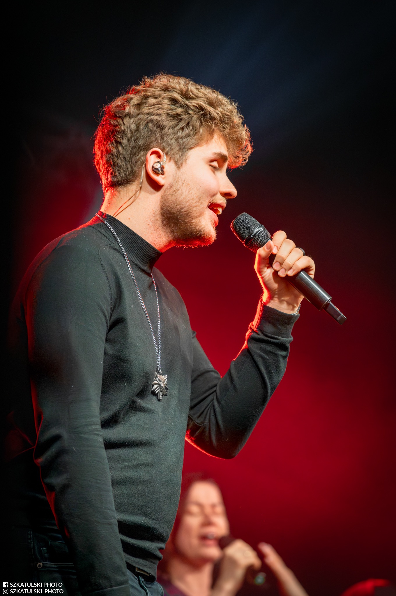 Piosenkarz z mikrofonem na scenie w czerwonym oświetleniu. Widoczny srebrny naszyjnik z symbolem wilka. W tle rozmyta postać kobiety.