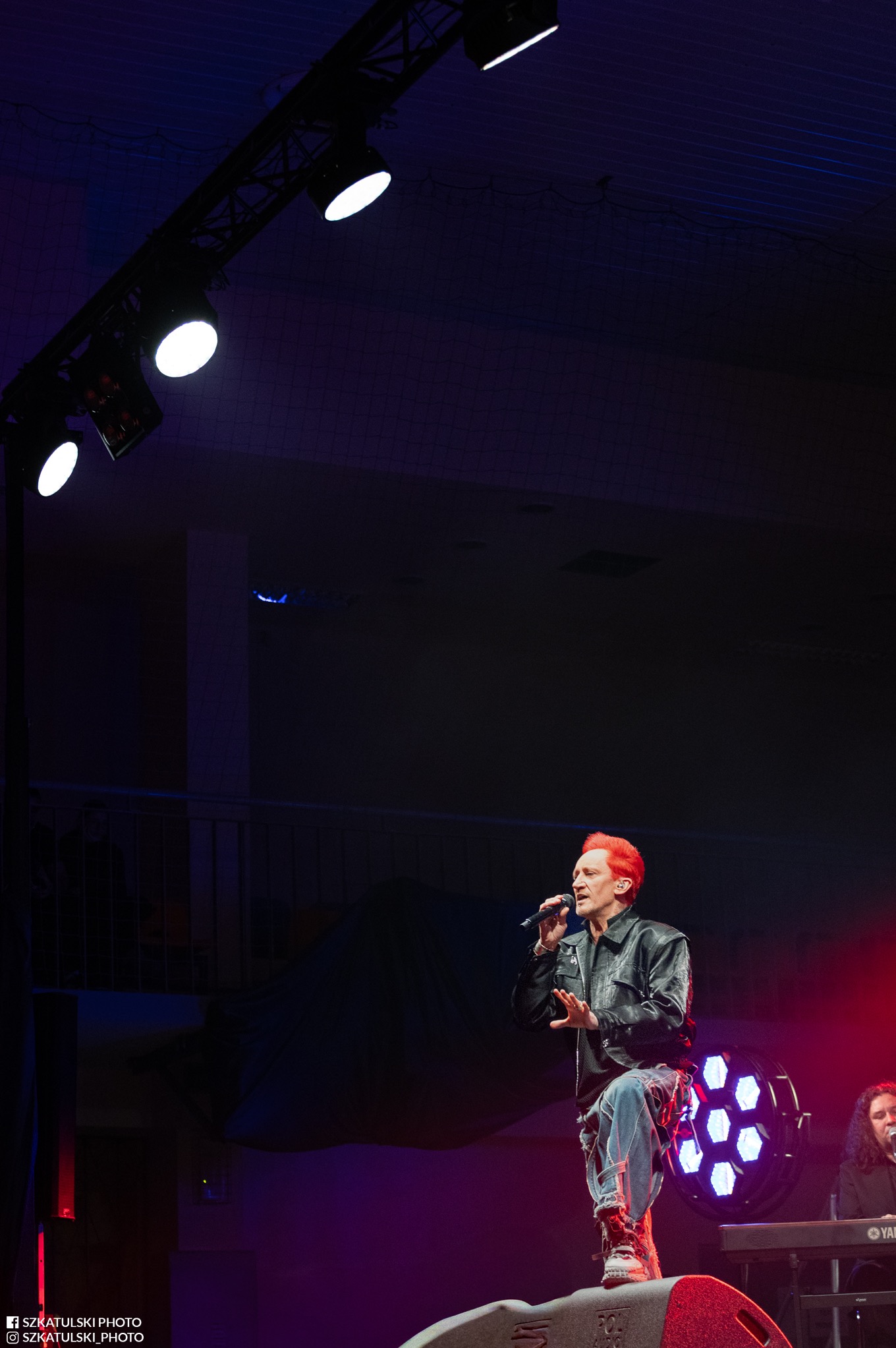 Wokalista z płomiennymi włosami w skórzanej kurtce, śpiewa do mikrofonu na scenie oświetlonej reflektorami. W tle widoczna klawiatura i fragment widowni.
