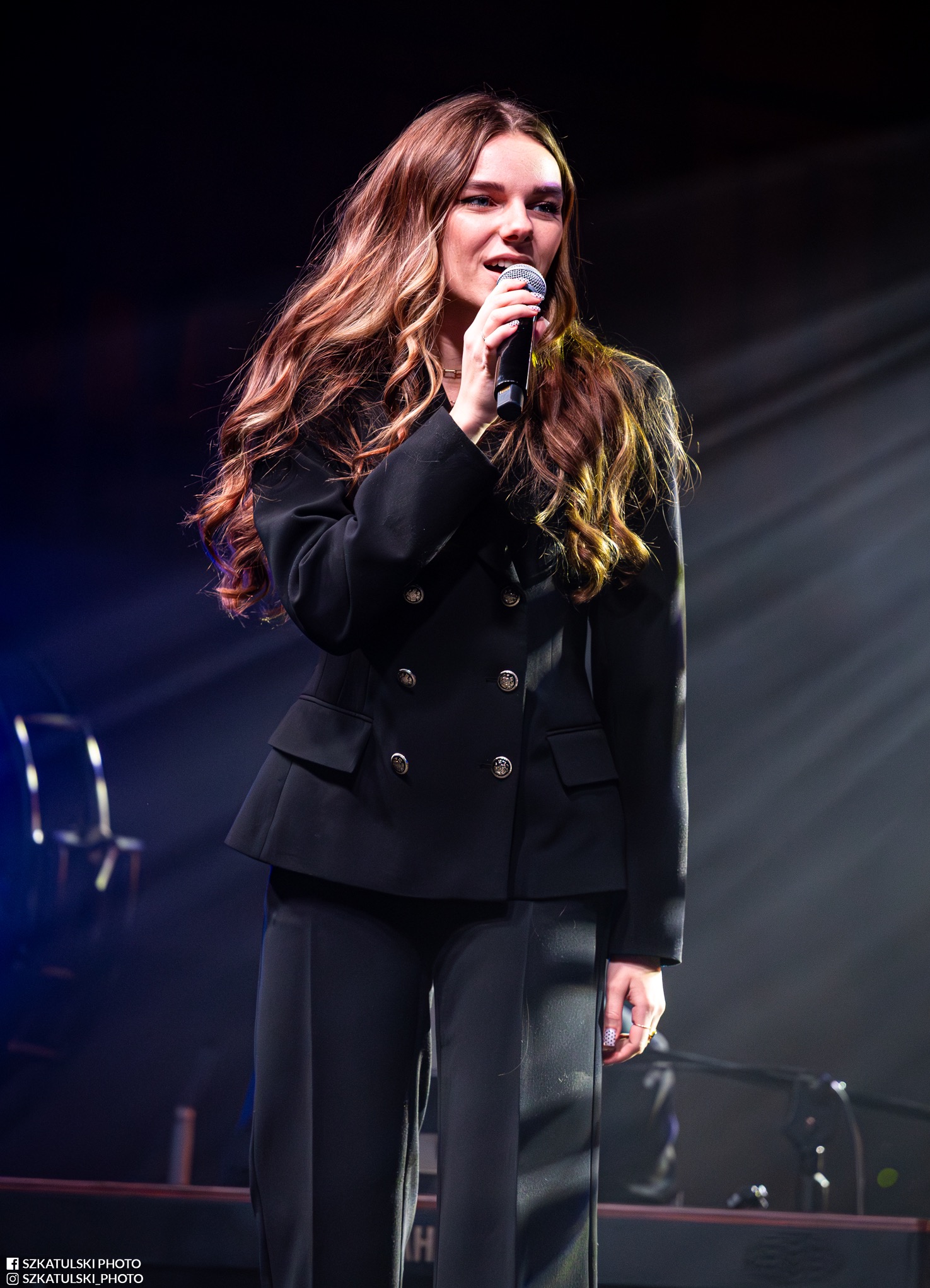 Piosenkarka w czarnym garniturze, z mikrofonem na scenie podczas występu. Długie, falowane włosy, profesjonalny makijaż. Scena oświetlona reflektorami.