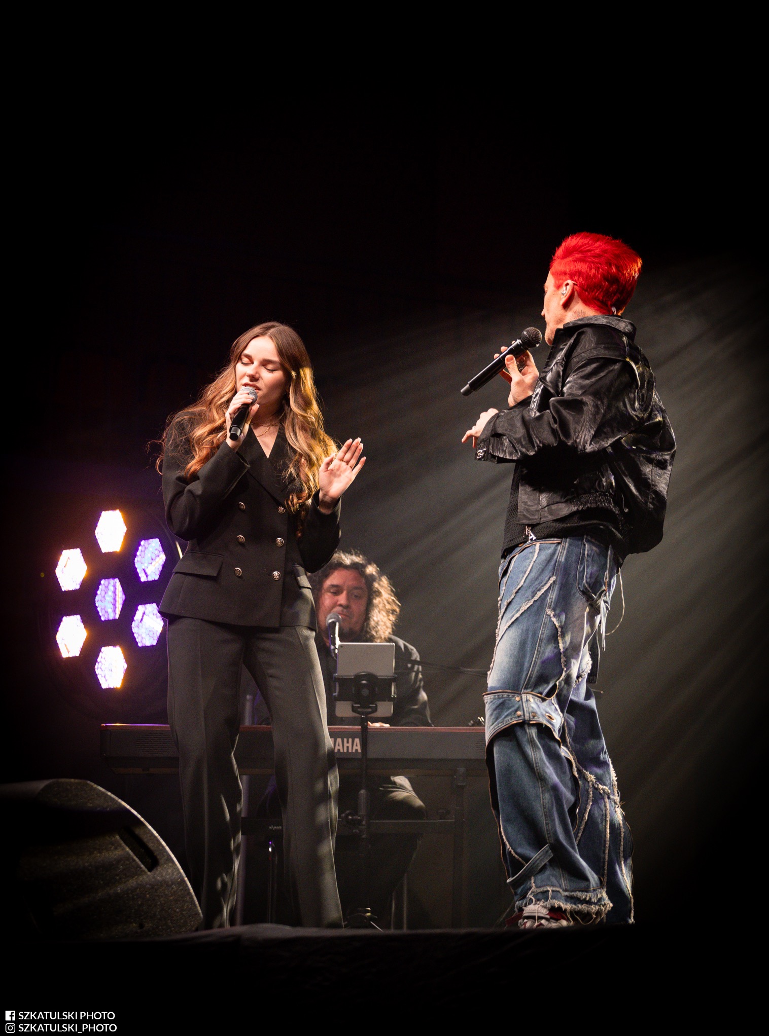 Dwoje wokalistów na scenie podczas koncertu. Kobieta w garniturze i mężczyzna z czerwonymi włosami śpiewają do mikrofonów. W tle klawiszowiec i oświetlenie sceniczne.