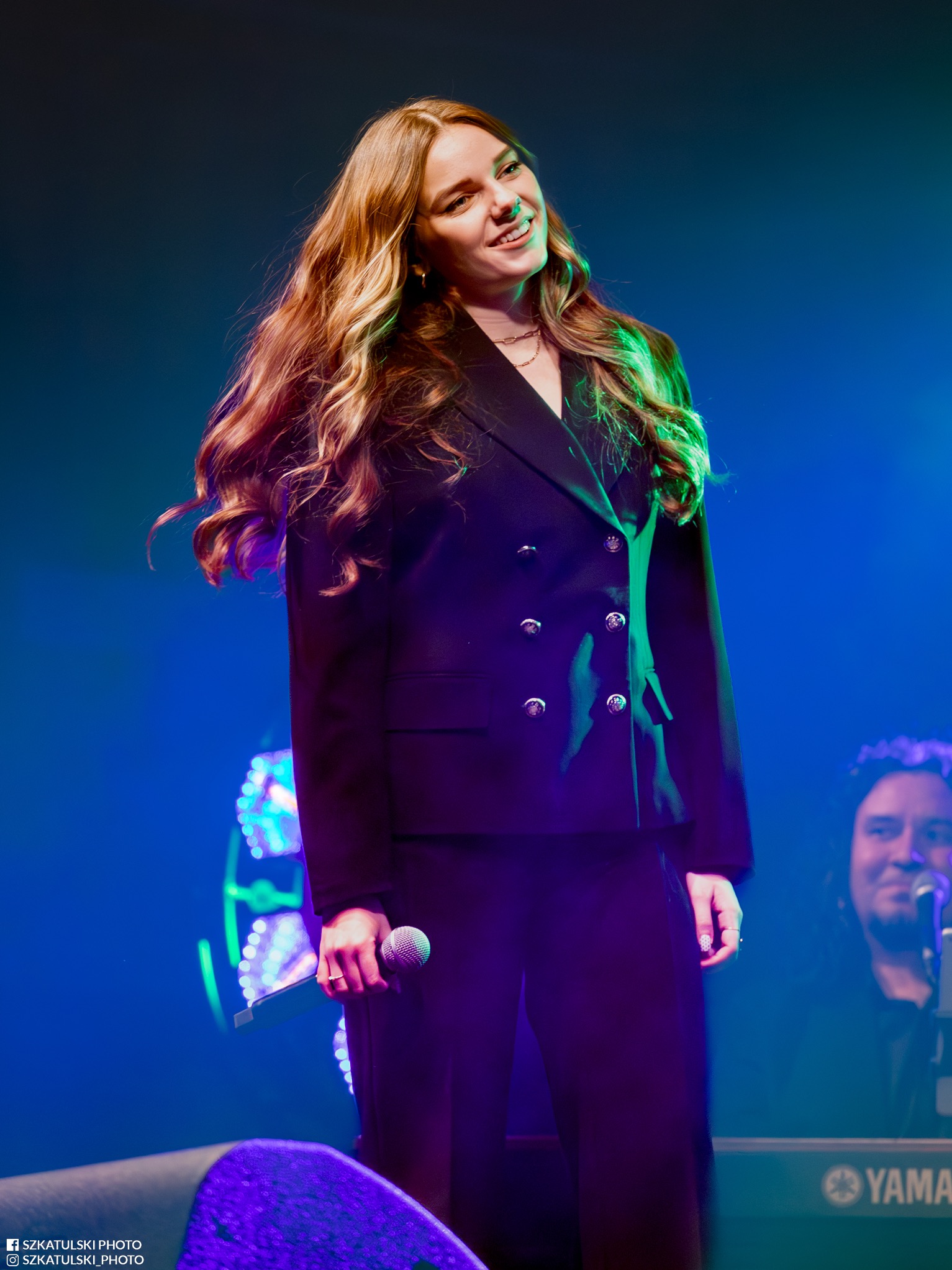 Uśmiechnięta wokalistka w czarnym garniturze, trzymająca mikrofon na scenie z niebieskim oświetleniem i klawiszowcem w tle. Koncertowy kadr.