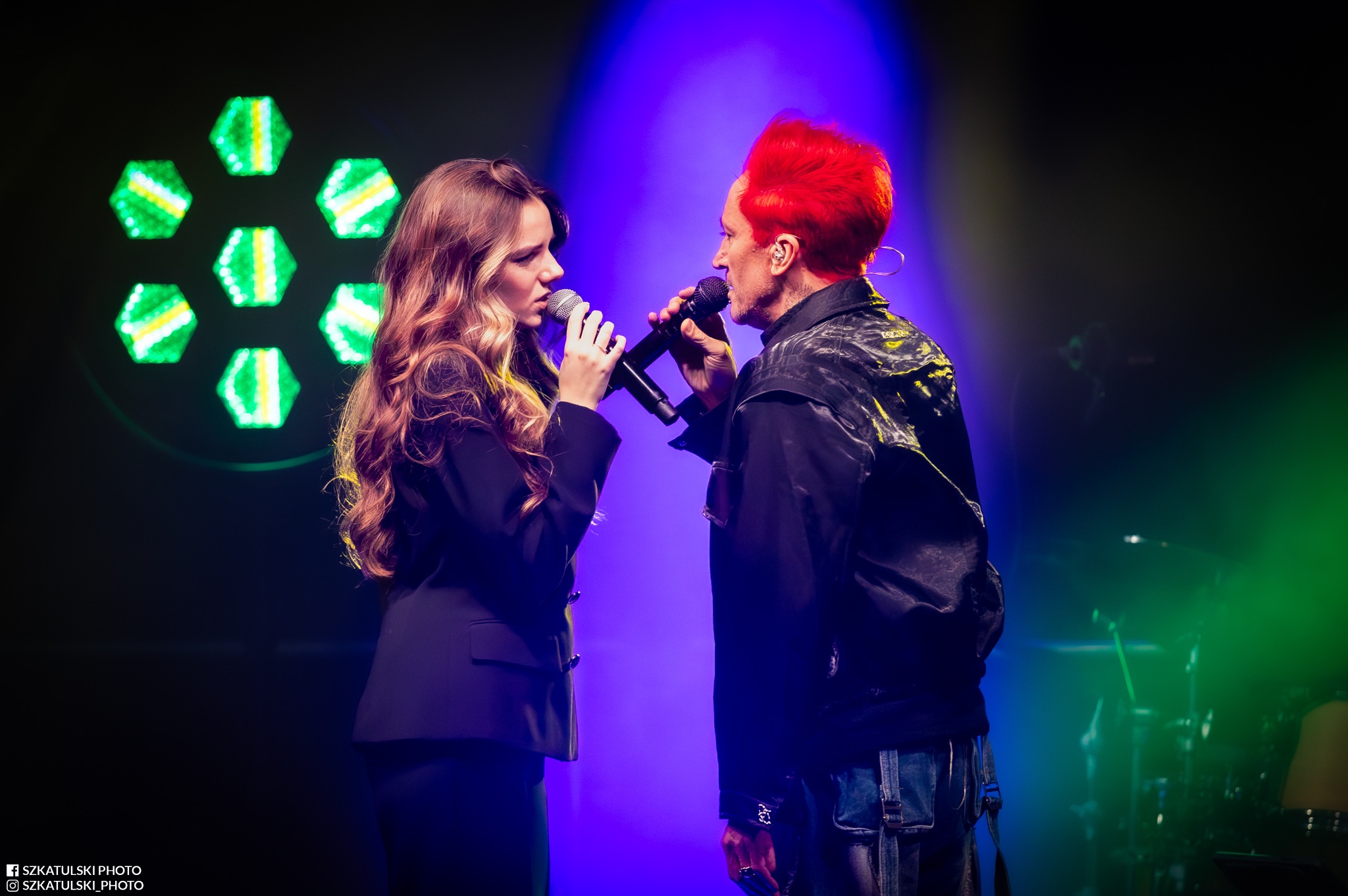 Duet wokalny na scenie w Sulejowie: kobieta w czarnym żakiecie i mężczyzna z czerwonymi włosami śpiewają do mikrofonów w blasku kolorowych świateł.