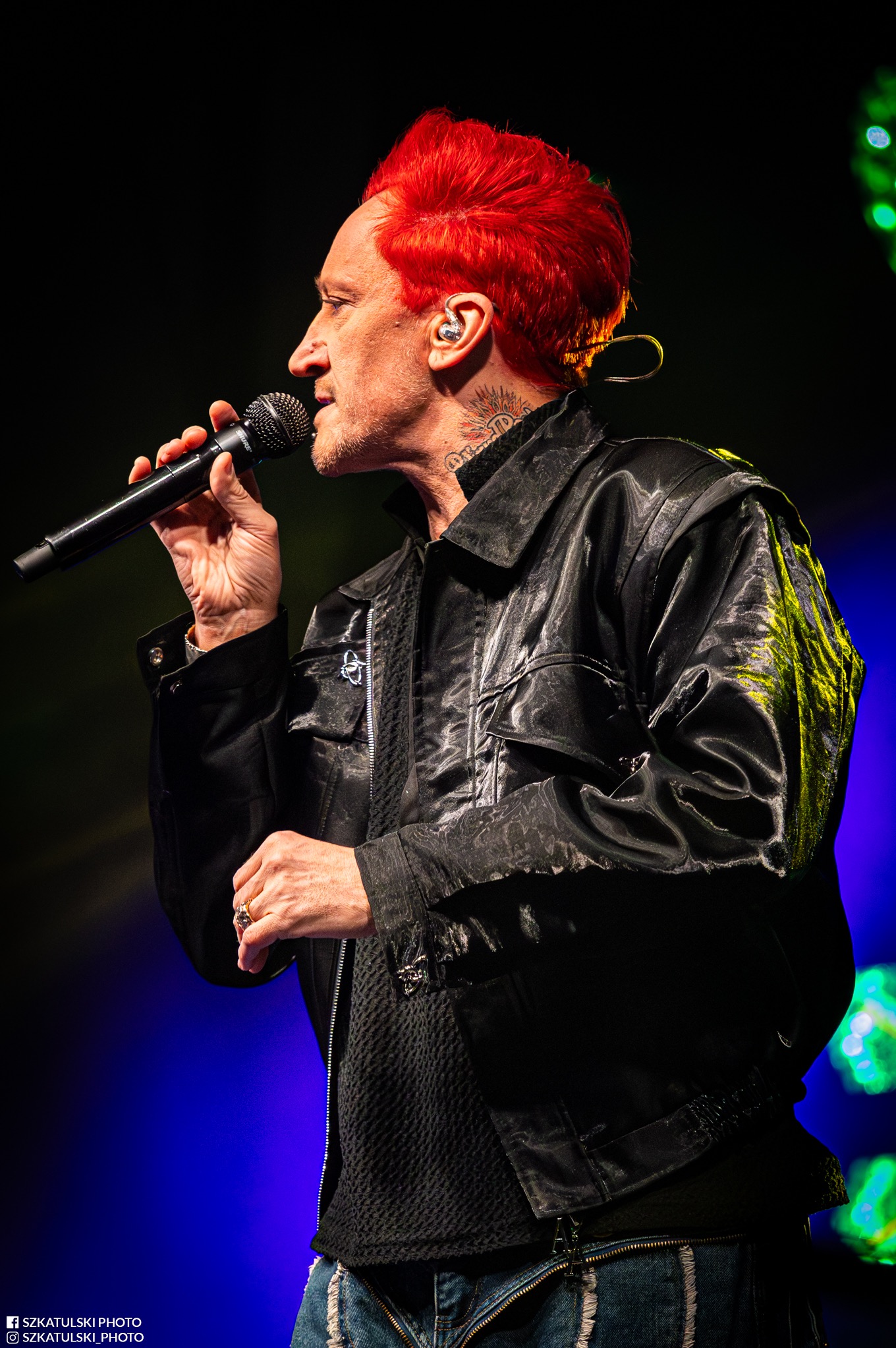 Profil wokalisty z czerwonymi włosami, śpiewającego do mikrofonu na scenie. Widoczne tatuaże na szyi, błyszcząca kurtka i dżinsy. Scena oświetlona niebieskim i zielonym światłem.