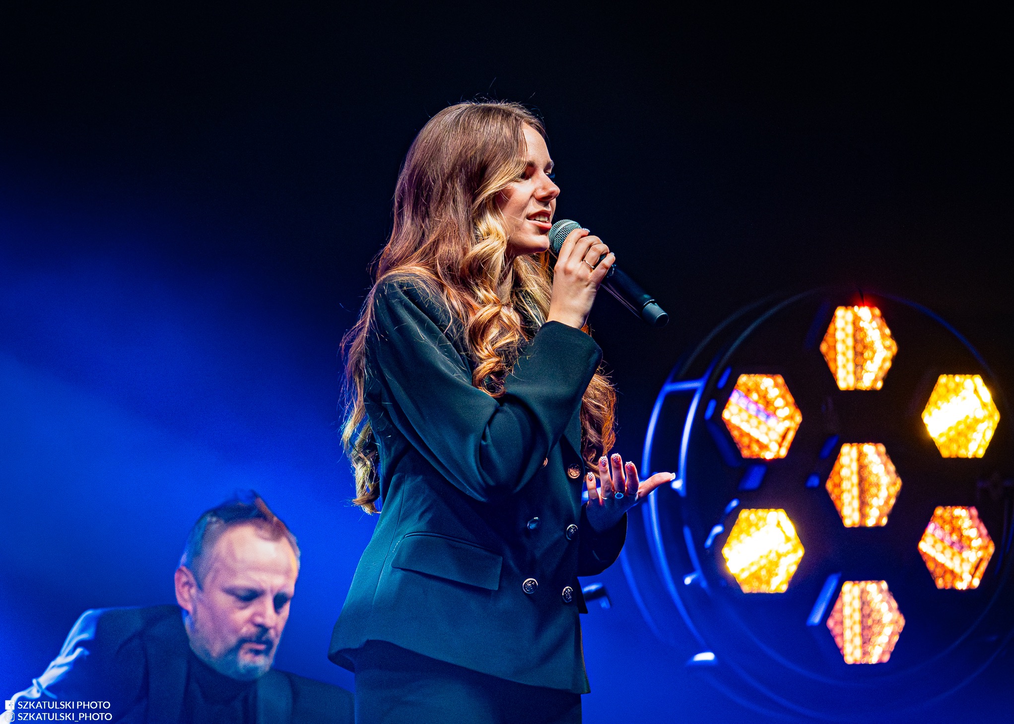 Piosenkarka w czarnym garniturze śpiewa do mikrofonu na scenie z oświetleniem. W tle muzyk. Ujęcie z dołu, podkreślające energię występu.
