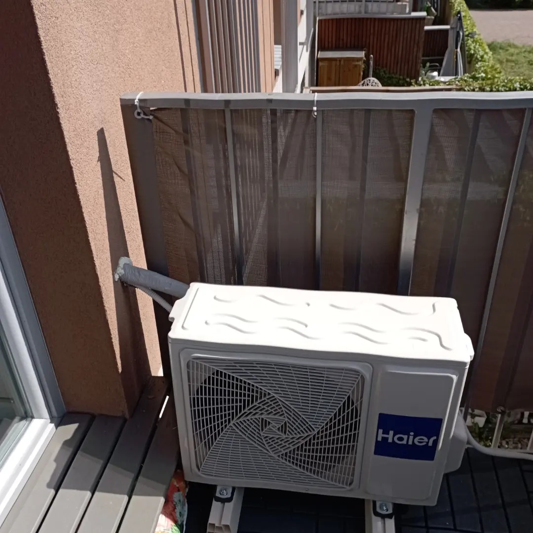 Zewnętrzna jednostka klimatyzacji Haier zamontowana na balkonie, widoczna z boku, z szarą osłoną balkonu w tle i fragmentem elewacji budynku.