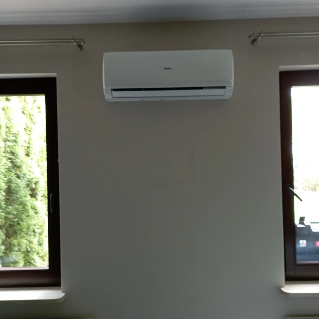 Biały klimatyzator Haier zamontowany na szarej ścianie między dwoma oknami z ciemnymi ramami. Widok na zieleń za oknem.