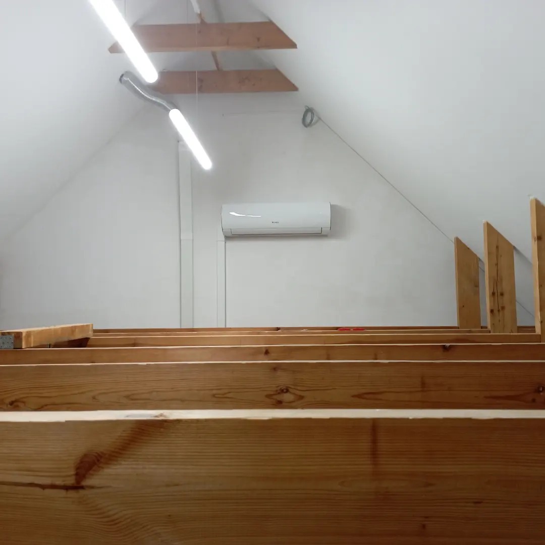 Biały klimatyzator na ścianie poddasza z drewnianą konstrukcją stropu. Widok z dołu, oświetlenie jarzeniowe.
