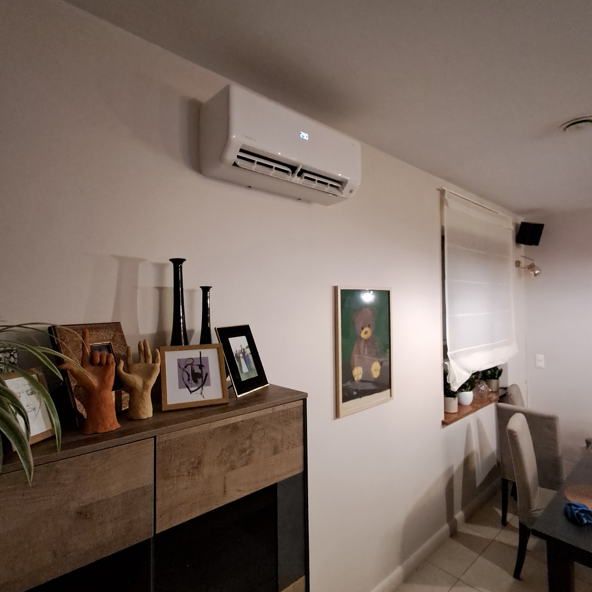 Biała klimatyzacja ścienna z wyświetlaczem temperatury w salonie, nad komodą z dekoracjami i obrazami. Widok na okno z roletą rzymską.