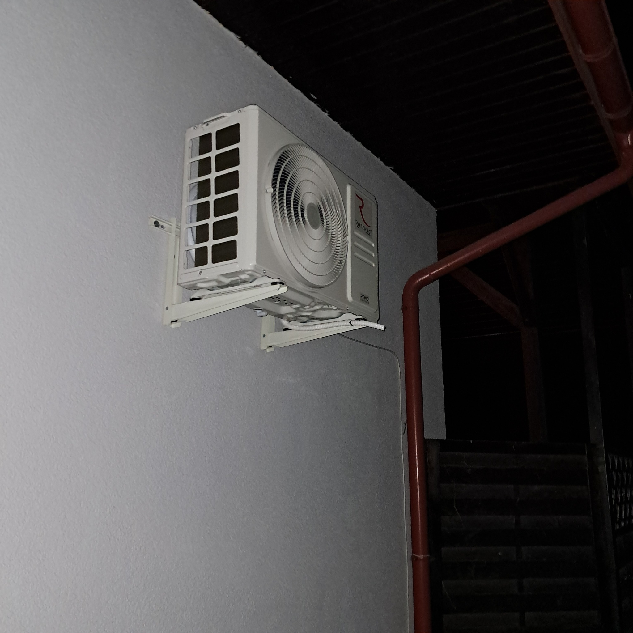 Biały klimatyzator Rotenso zamontowany na szarej ścianie zewnętrznej budynku, widoczny fragment rynny. Ujęcie z dołu, wieczorne oświetlenie.