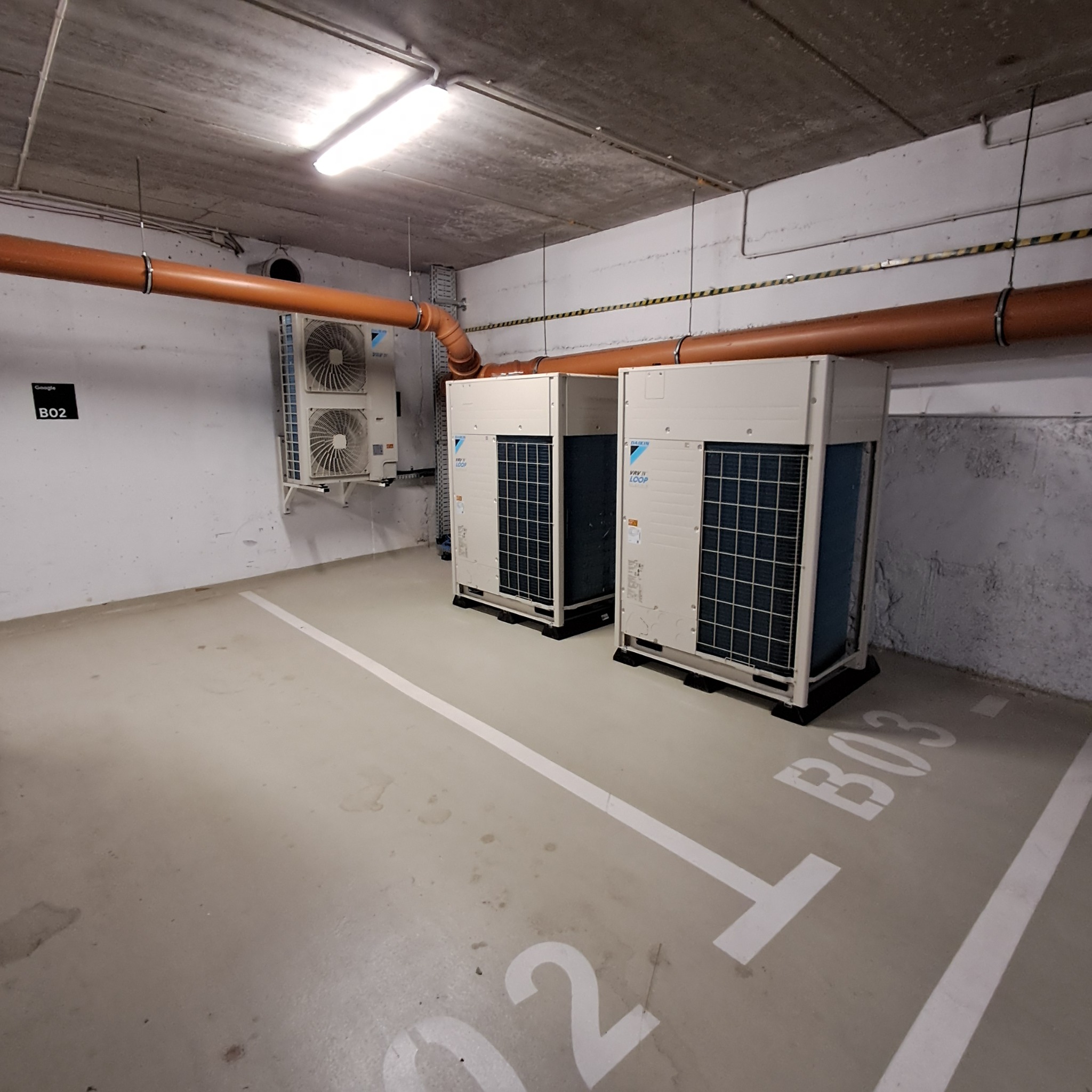 Dwa agregaty klimatyzacyjne Daikin VRV IV Loop w garażu podziemnym, z widoczną instalacją rurową pod sufitem i oznaczeniami miejsc parkingowych. Szare tło ścian i sufitu.