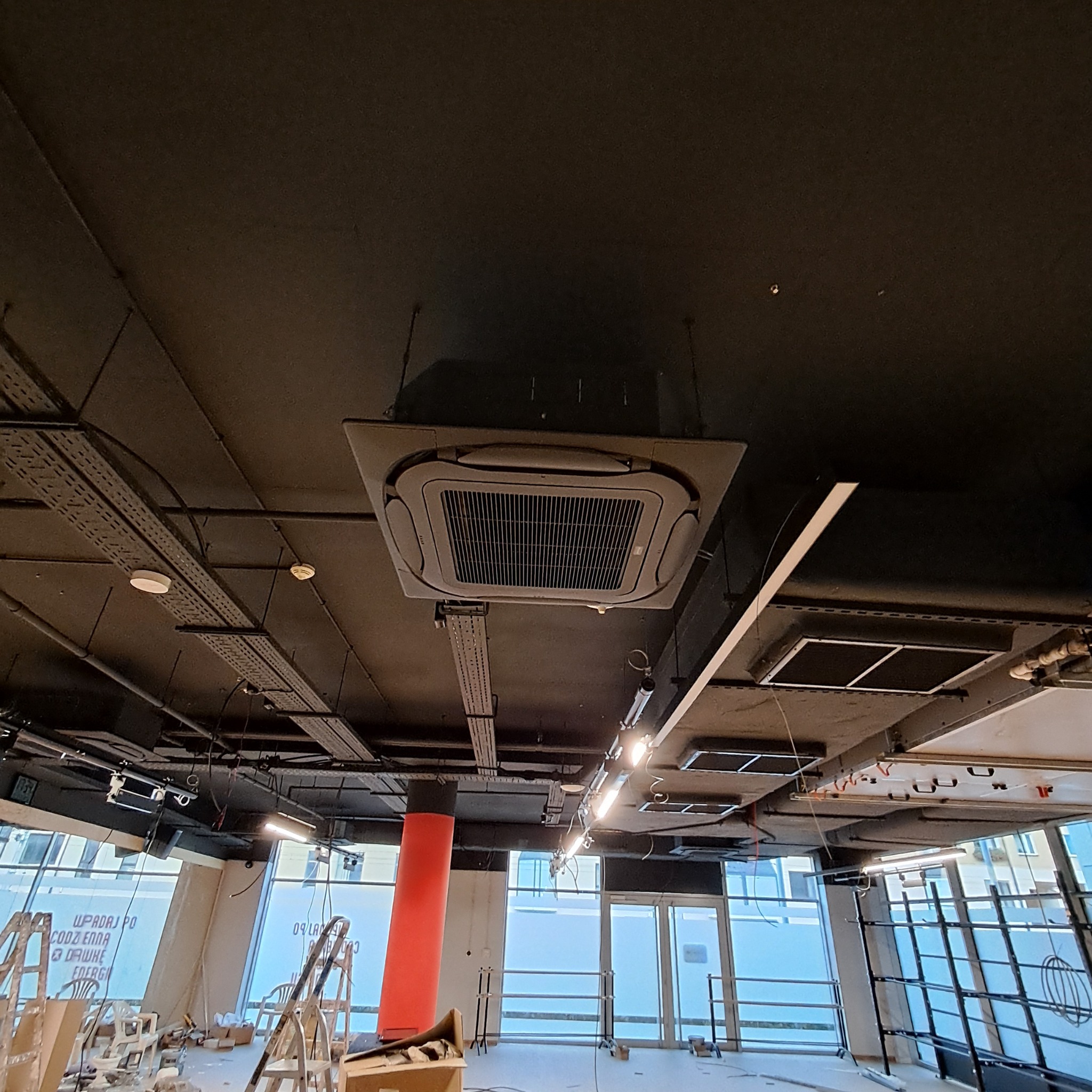 Kasetonowa klimatyzacja sufitowa w biurze, widok z dołu. Widoczne instalacje, kable i elementy konstrukcyjne sufitu. W tle okna i elementy wyposażenia.