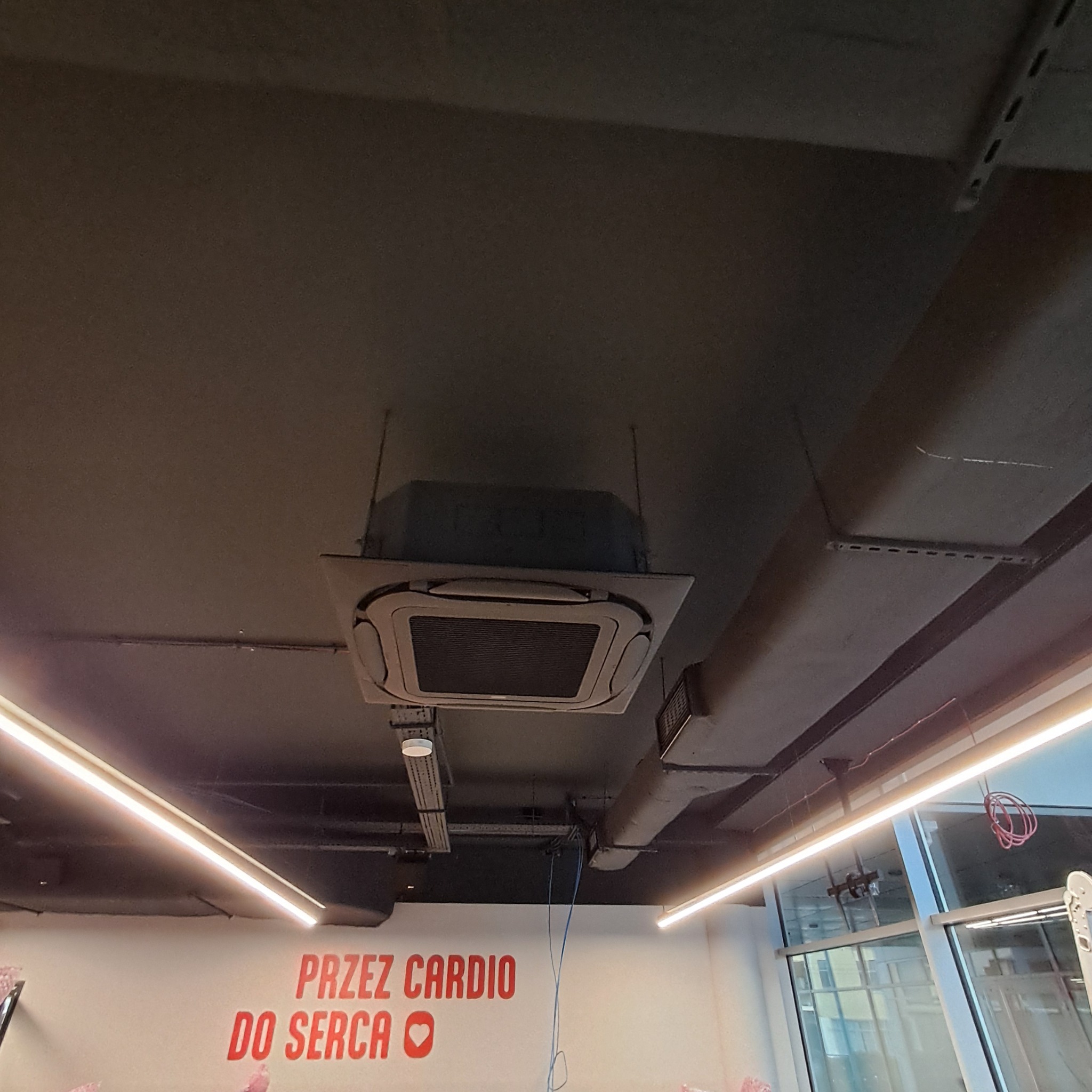 Sufitowa klimatyzacja kasetonowa w pomieszczeniu komercyjnym, widoczne przewody i oświetlenie LED. Ujęcie z dołu, ciemna kolorystyka sufitu.