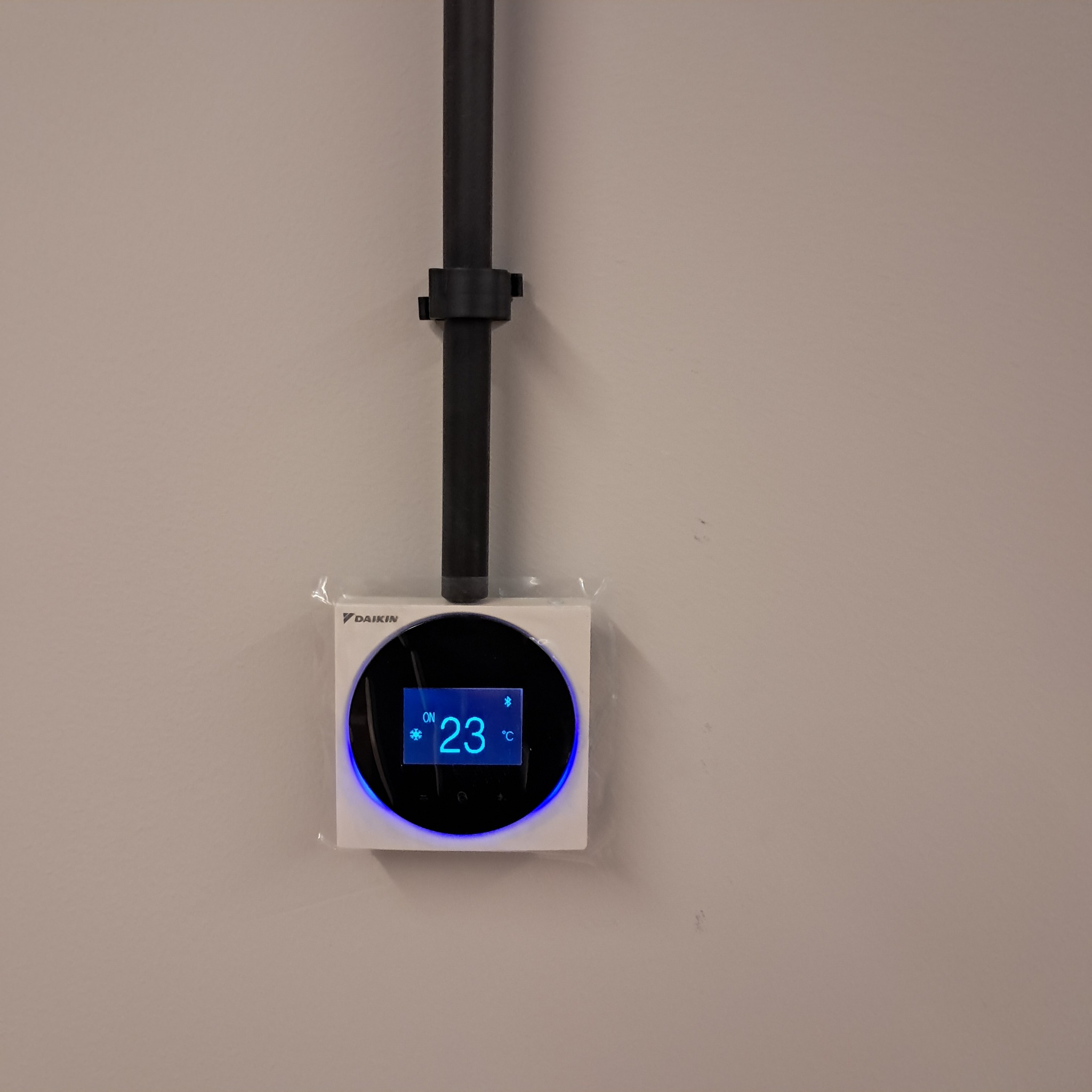 Nowoczesny, biały panel sterowania klimatyzacją Daikin z czarnym, okrągłym wyświetlaczem, zamontowany na ścianie. Widoczna rura instalacyjna. Temperatura ustawiona na 23°C.