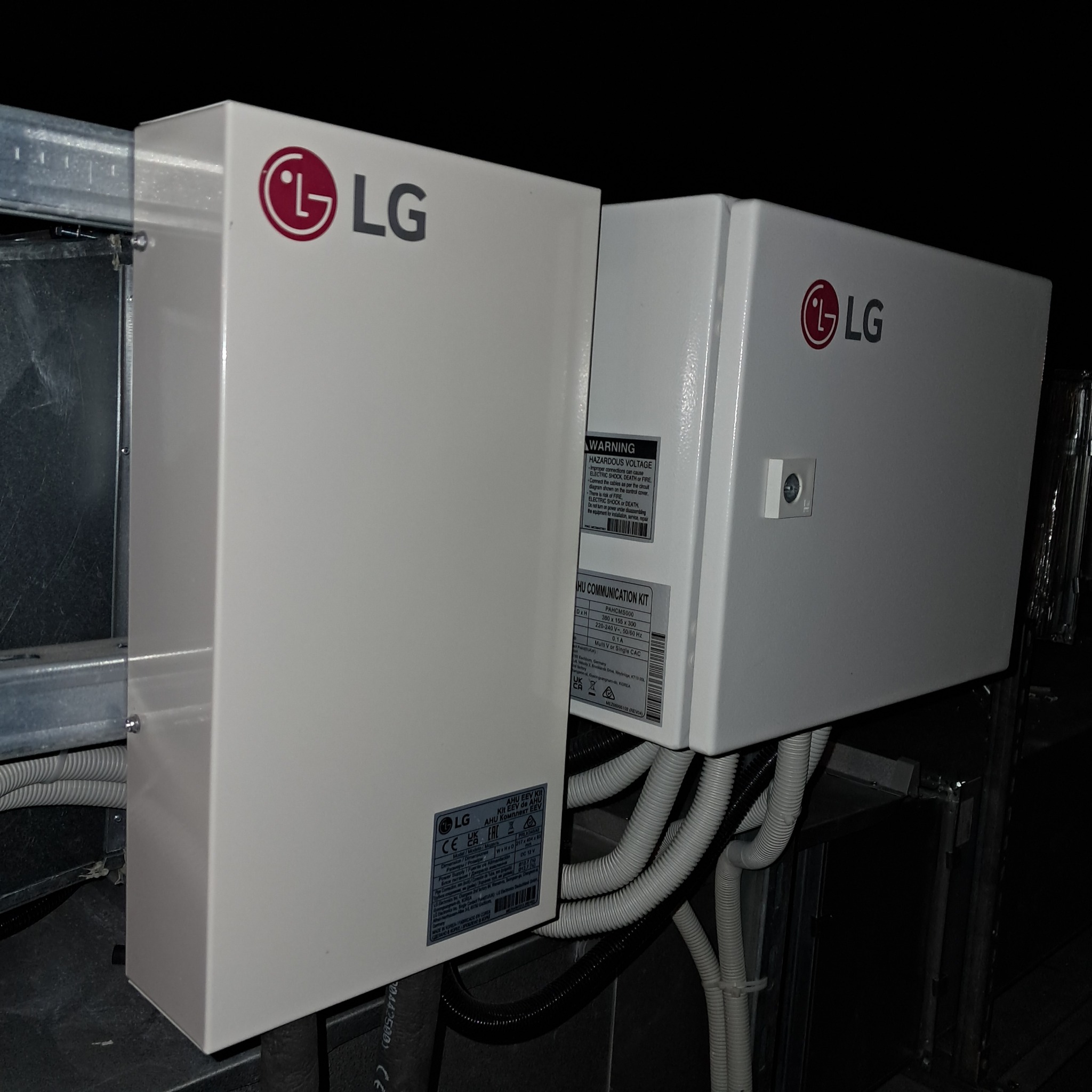 Dwa białe moduły klimatyzacji LG z widocznymi przewodami, zamontowane na metalowej konstrukcji. Na obudowach etykiety informacyjne i logo producenta.