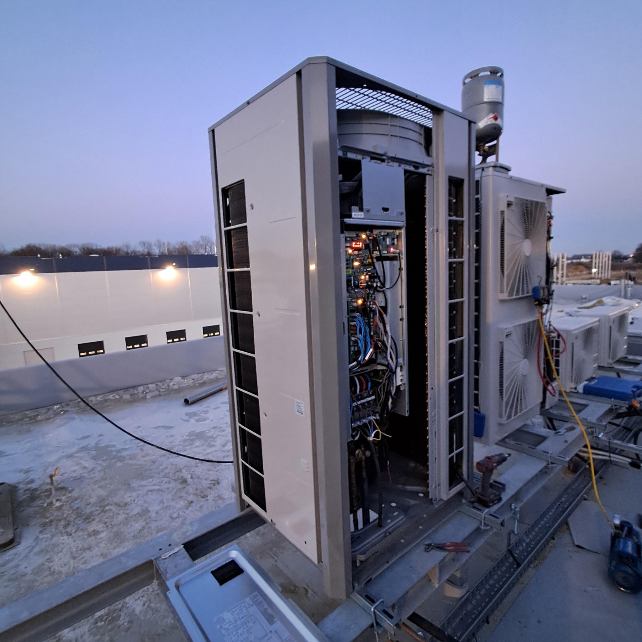 Instalacja klimatyzacji na dachu budynku. Otwarta obudowa z widocznymi przewodami i elektroniką. W tle inne jednostki klimatyzacyjne i budynek przemysłowy.