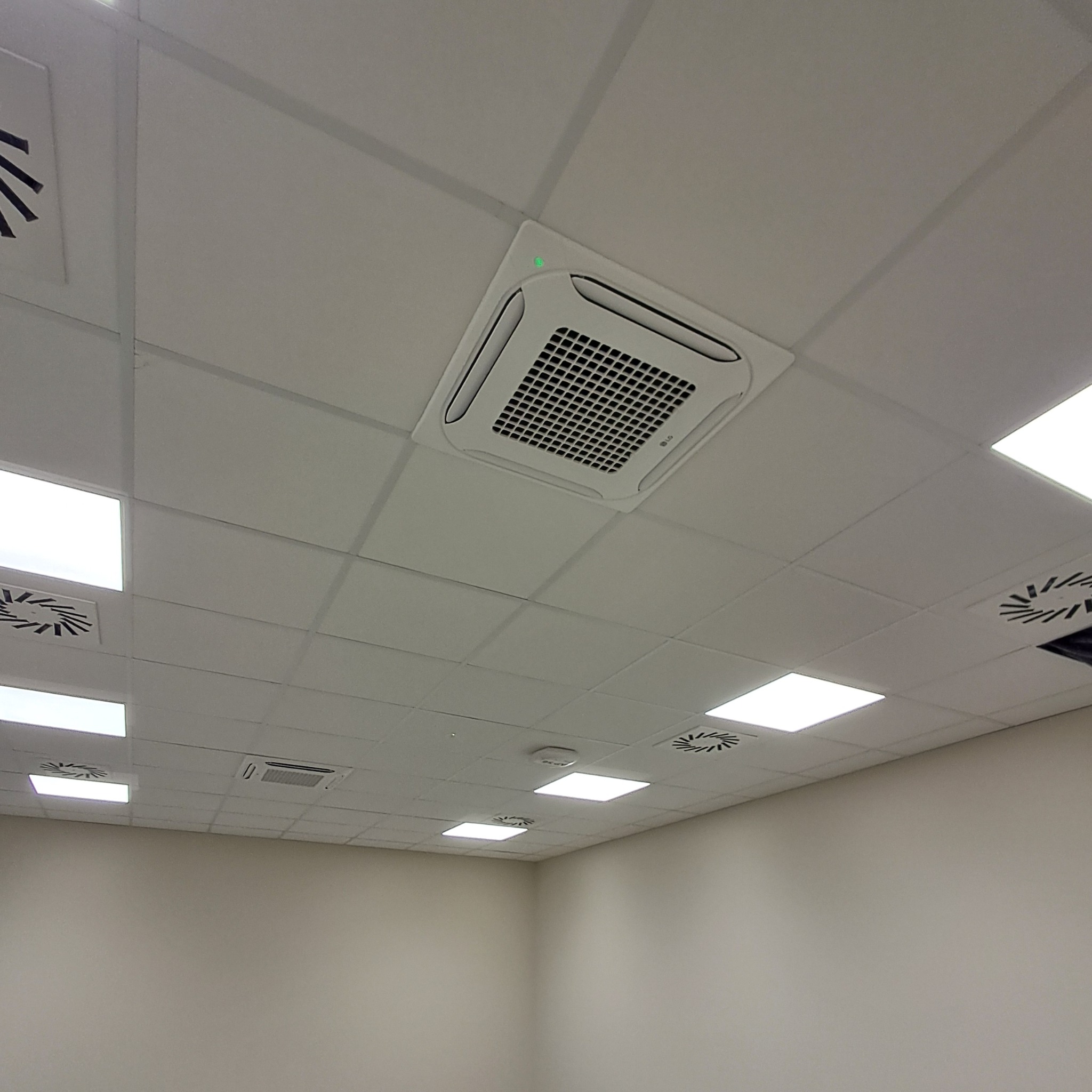 Kasetonowa klimatyzacja LG wkomponowana w sufit podwieszany z oświetleniem LED. Widok z dołu na białe urządzenie z zieloną diodą sygnalizacyjną.