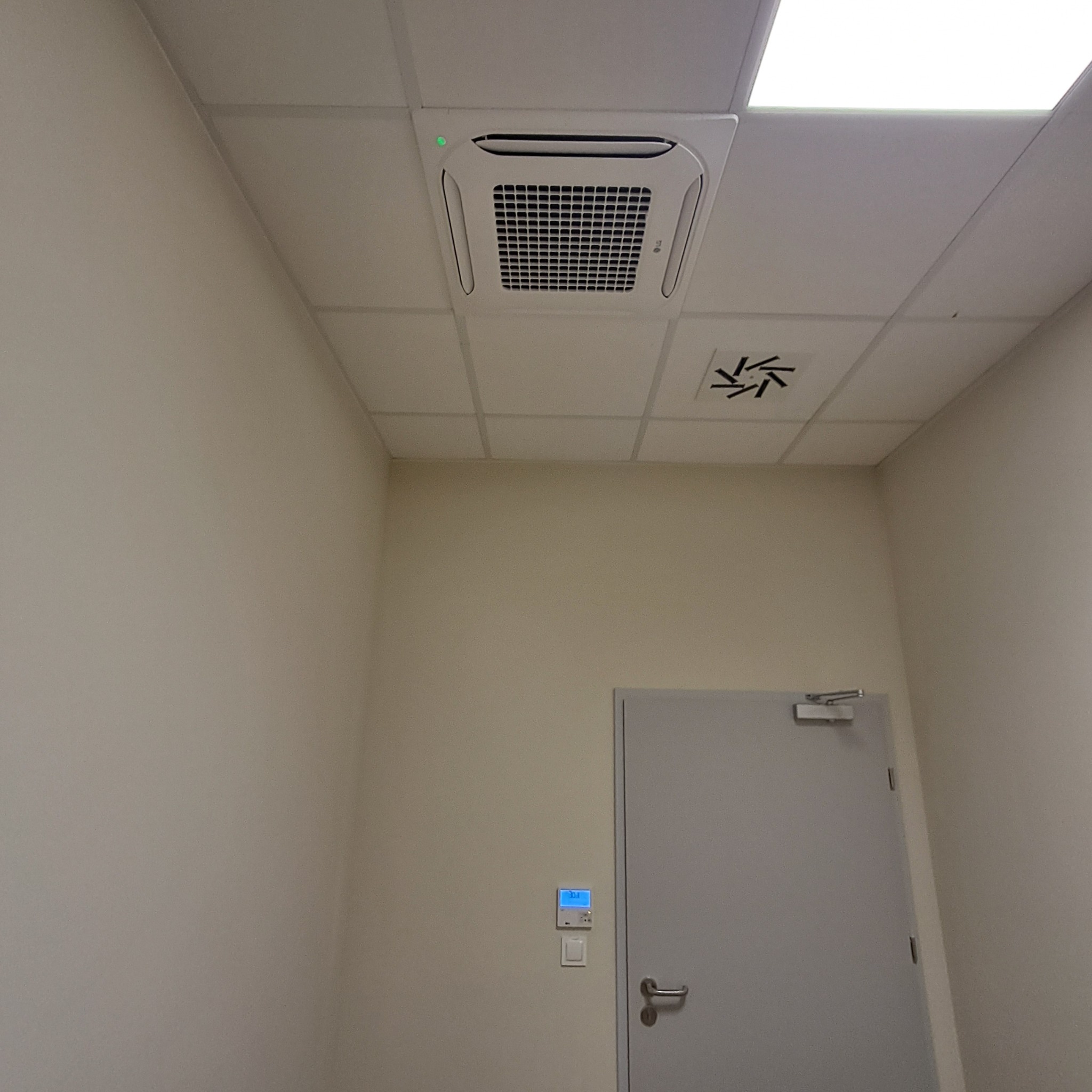 Biała, sufitowa kaseta klimatyzacyjna z kratką, zamontowana w biurze. Widoczny panel sterowania na ścianie i drzwi. Oświetlenie sufitowe.