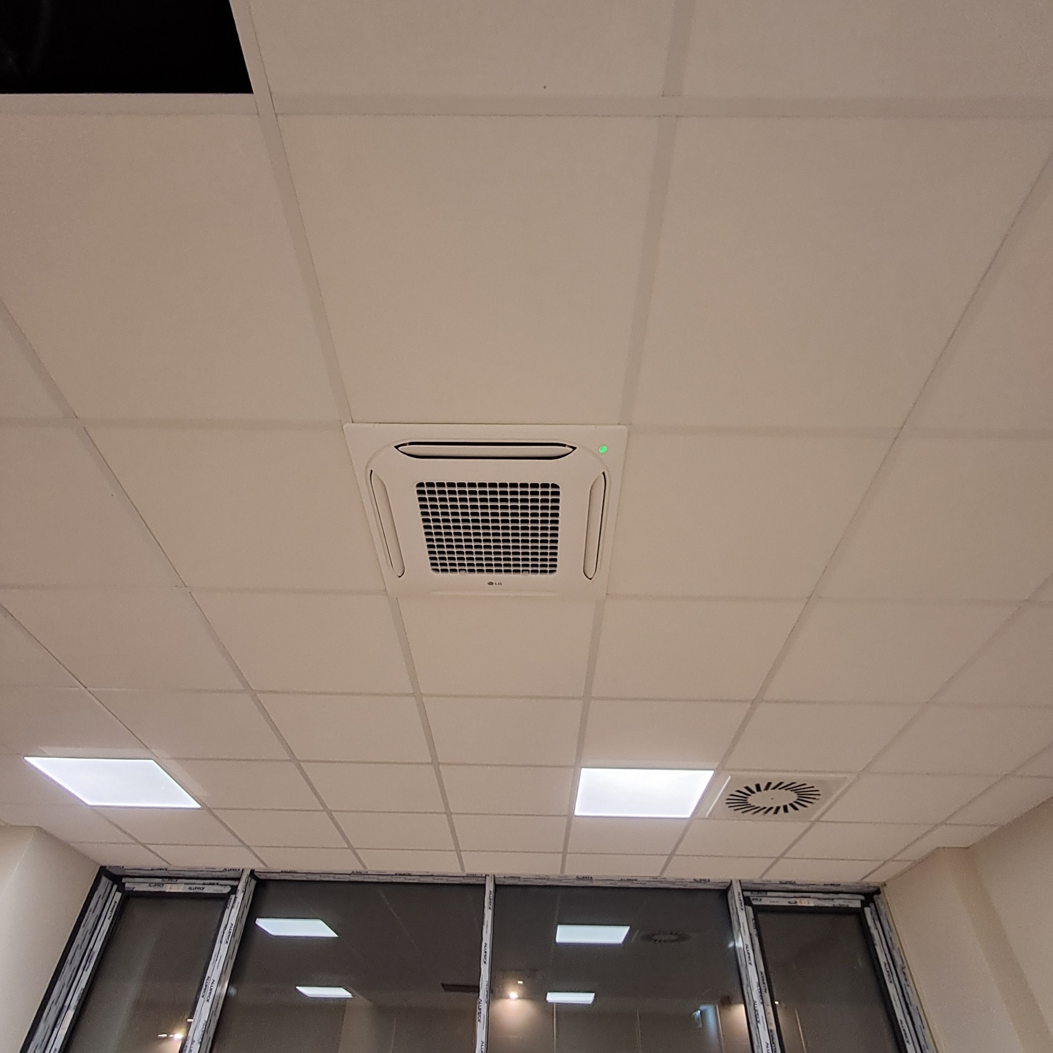 Kasetonowa klimatyzacja LG zamontowana w suficie podwieszanym biura, widok z dołu. Widoczne okna z aluminiowymi ramami i kratka wentylacyjna.
