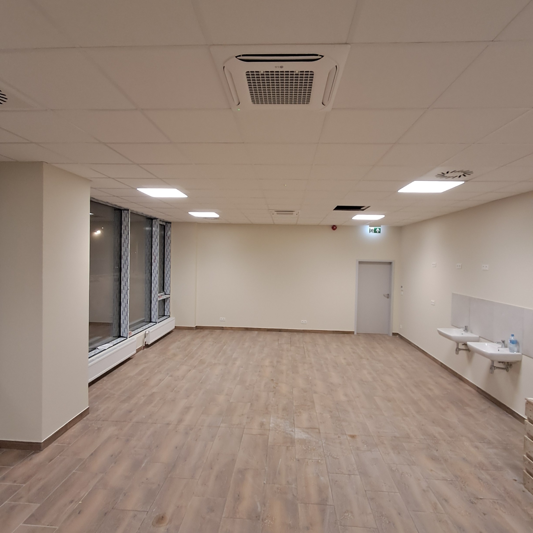 Klimatyzator kasetonowy LG zamontowany w suficie podwieszanym, widok na przestronne, jasne pomieszczenie z oknami i umywalkami. Wykończona instalacja w biurze.