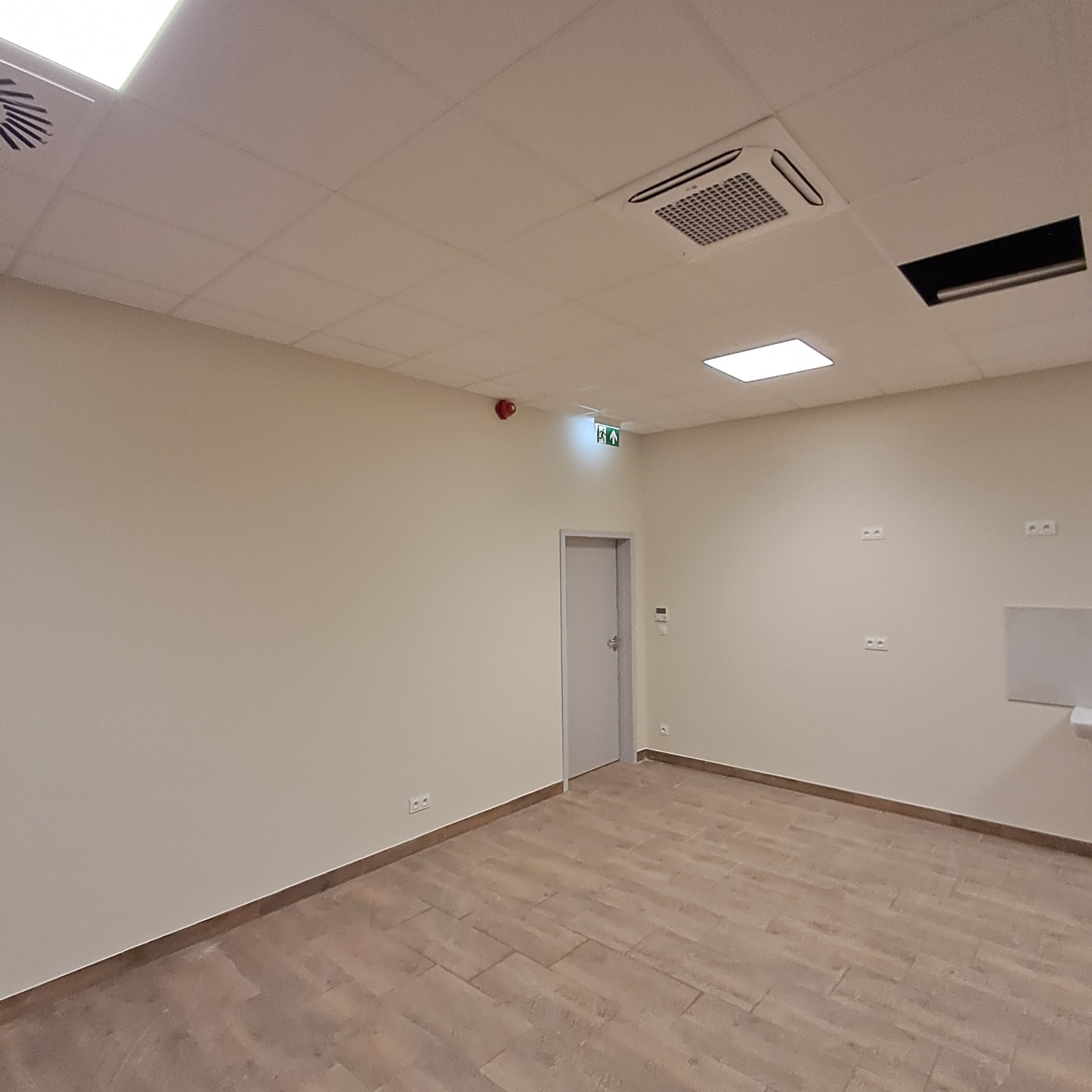 Wnętrze biura z zamontowaną klimatyzacją kasetonową w suficie podwieszanym. Widoczne gniazdka elektryczne, drzwi ewakuacyjne i fragment instalacji wentylacyjnej.