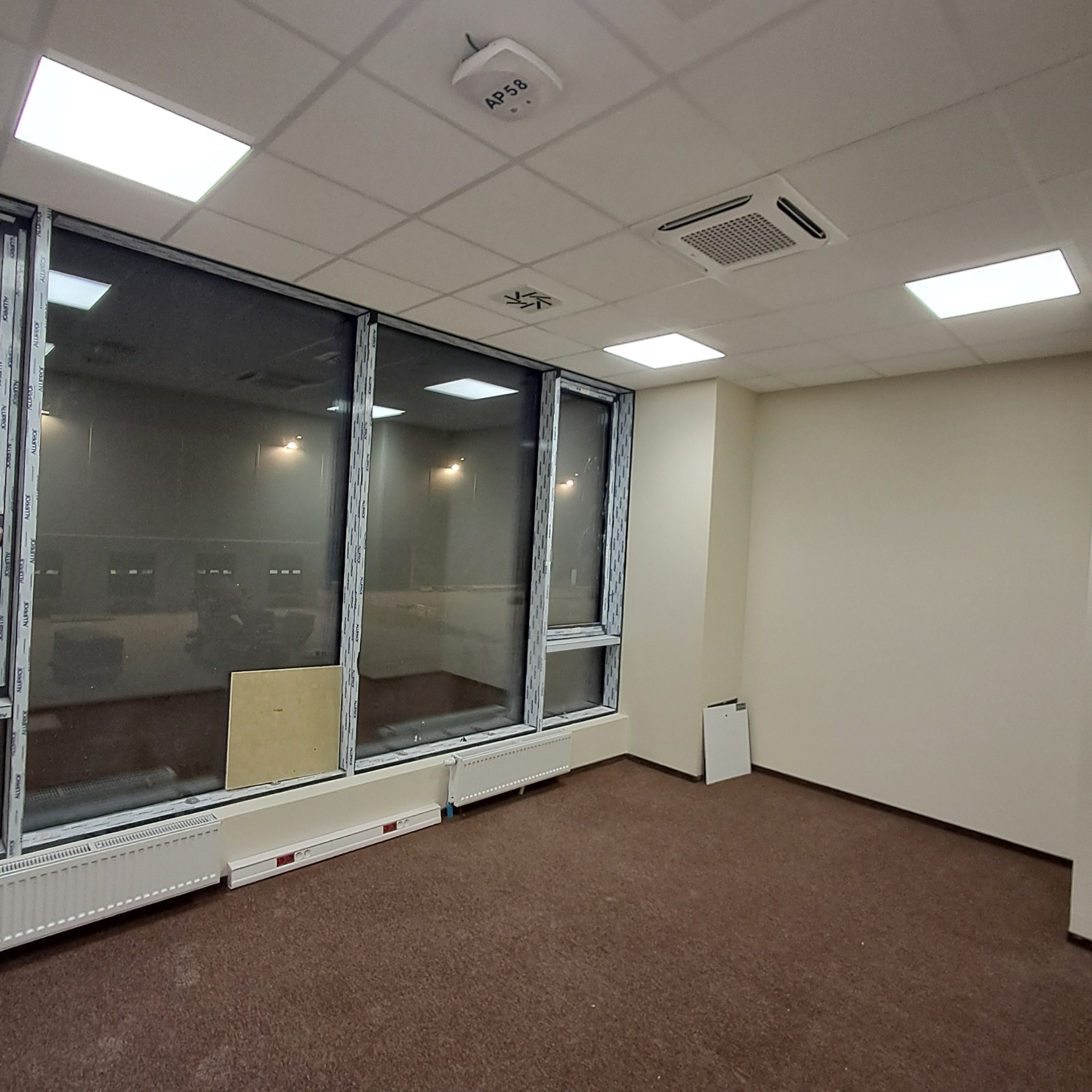 Wnętrze biura z dużymi oknami, grzejnikami pod oknami i zamontowaną klimatyzacją kasetonową w suficie. Widoczne elementy instalacji elektrycznej i wentylacyjnej.