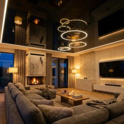 Elegancki sufit napinany w głębokim grafitowym odcieniu z subtelnym połyskiem, podkreślony ciepłą, ciągłą linią LED w kolorze kości słoniowej. Trzy efektowne, podwieszane pierścienie świetlne tworzą nowoczesną kompozycję