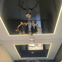 Powierzchnia odbija jak ciemne zwierciadło – widać w niej wyraźne odbicie złotego, industrialnego żyrandola w formie otwartego okręgu z widocznymi żarówkami i czarnymi linkami.