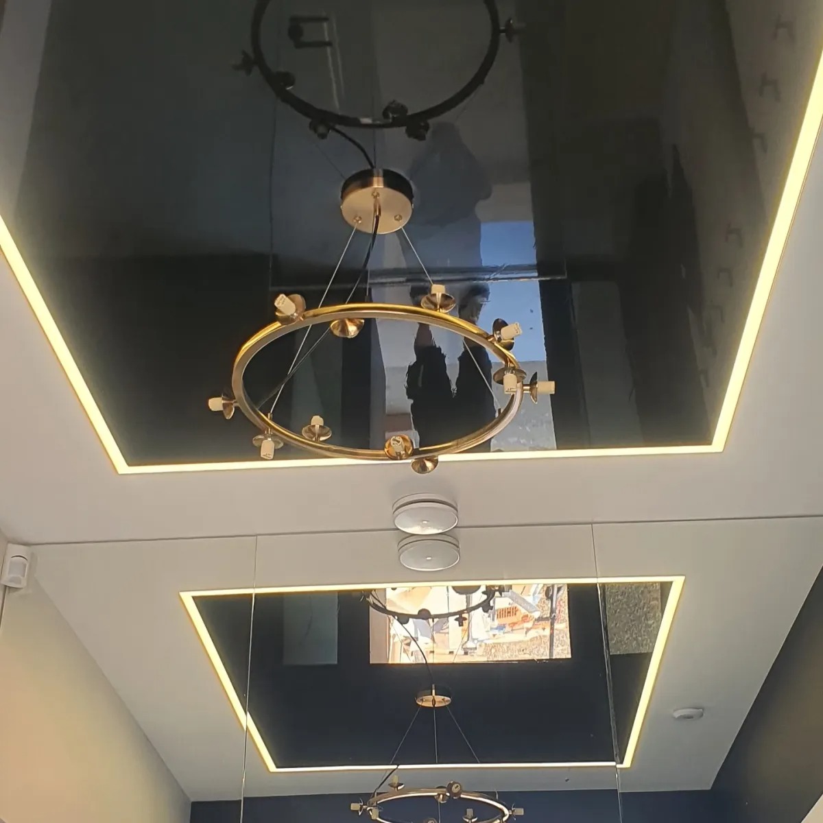 Błyszcząca, czarna powierzchnia sufitu odbija złoty, okrągły żyrandol z żarówkami. Sufit podświetlony listwami LED. Widoczne odbicie sylwetek.