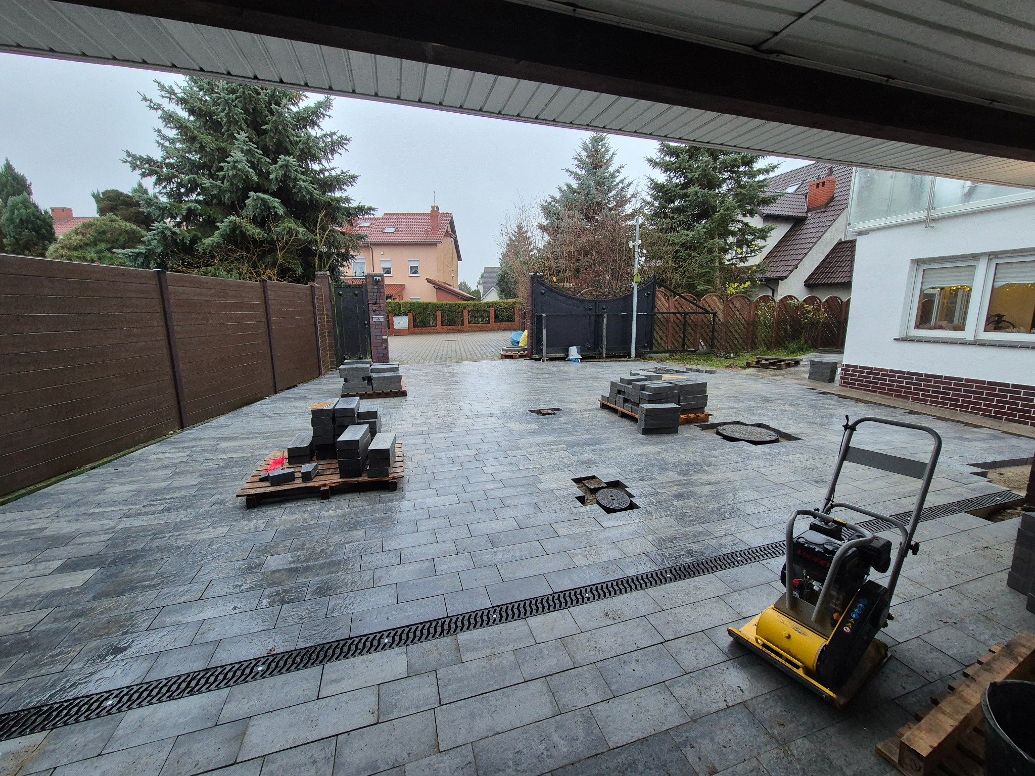 Układanie szarej kostki brukowej na podjeździe z widocznymi liniowymi odpływami. Na paletach kostka do ułożenia i zagęszczarka w trakcie prac.