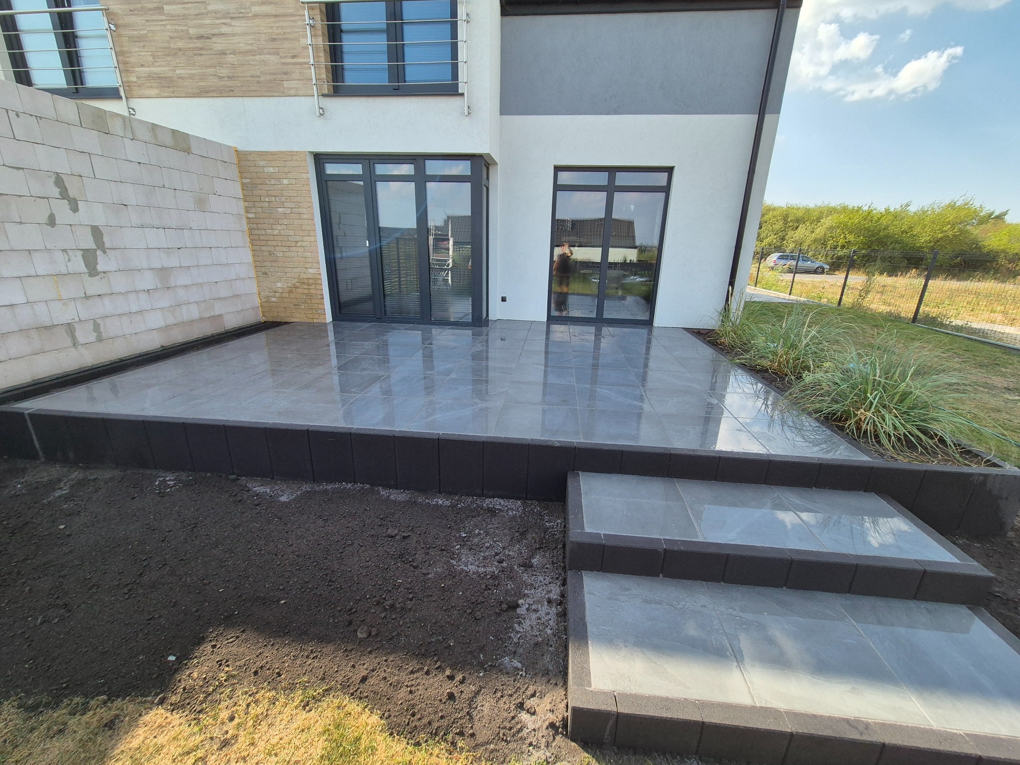 Nowoczesny taras z szarych płyt, obrzeża z ciemnej kostki, schody z tego samego materiału. Widok na dom z dużymi oknami i fragment ogrodu z zielenią.