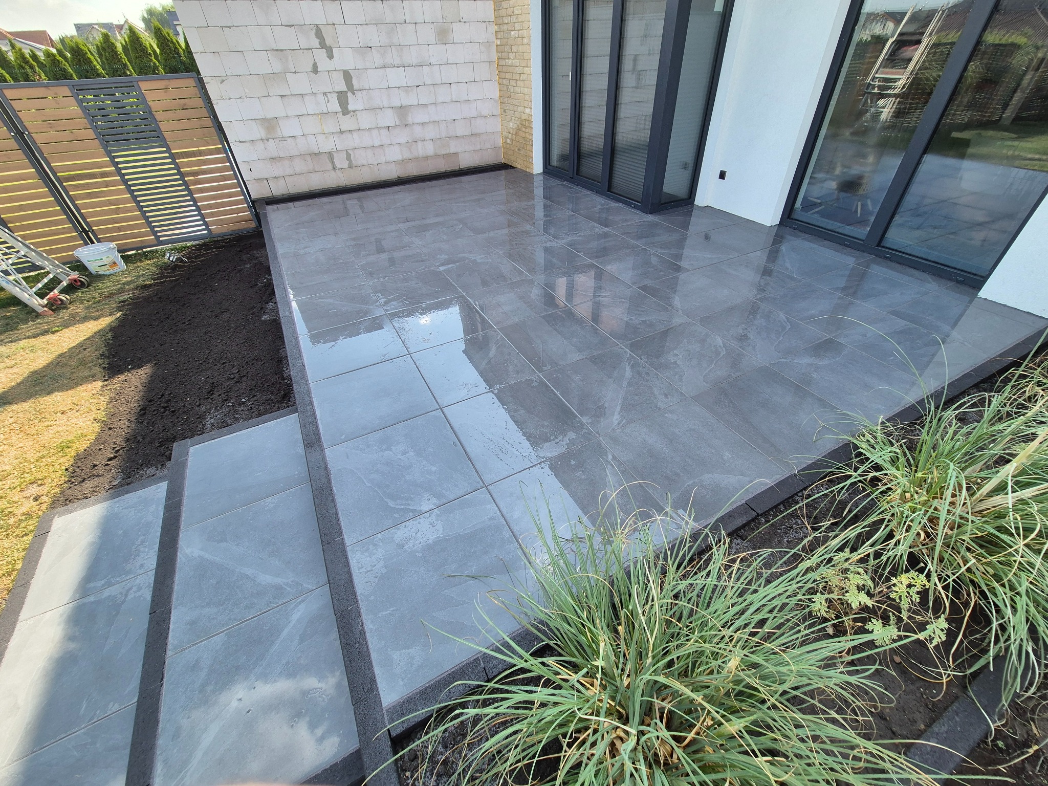 Nowoczesny taras z szarych płyt, z widocznymi schodami i fragmentem rabaty z trawami ozdobnymi. Minimalistyczny design, świeżo po deszczu, z odbiciami w mokrej powierzchni.
