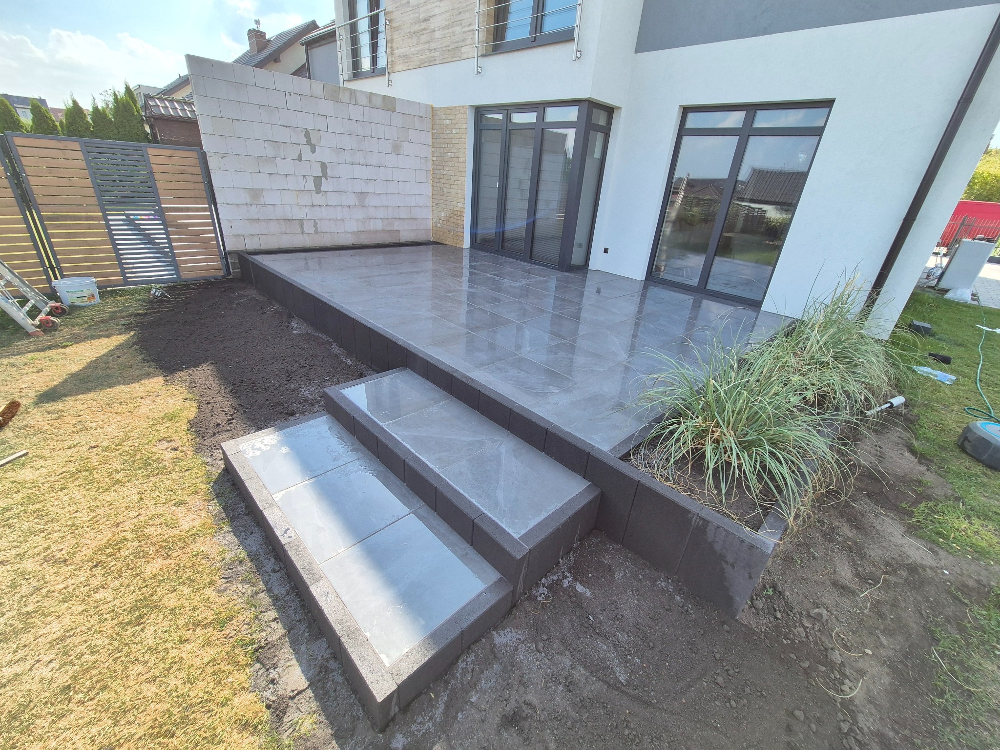 Nowoczesny taras z szarych płyt, z dwustopniowymi schodami i rabatą z trawą ozdobną. Widoczne elementy wykończeniowe i przygotowany teren pod dalsze prace.