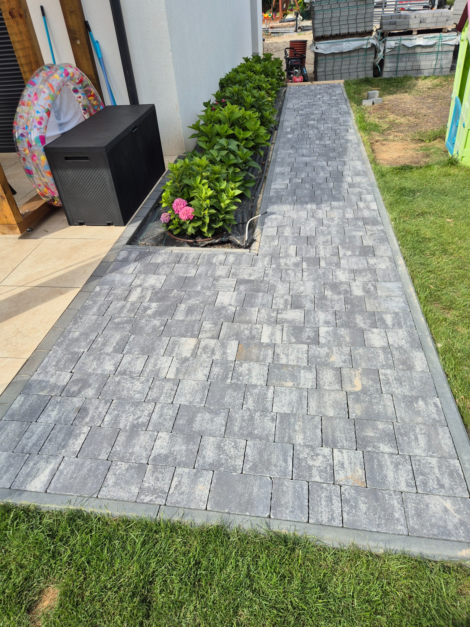 Ułożona ścieżka z szarej kostki brukowej, obrzeża z kostki, rabata z zielenią i różowymi kwiatami, trawnik. Widok z góry, słoneczny dzień.