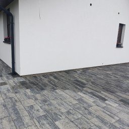 IZO-BAU Piotr Powroźnik - Biała elewacja domu z oknem i kwiatami, widok z poziomu kostki brukowej. Minimalistyczny design, ciemna rynna, nowoczesne wykończenie.