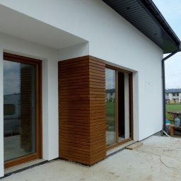 IZO-BAU Piotr Powroźnik - Biała elewacja domu z drewnianą okładziną narożną i oknami z brązowymi ramami. Widok z zewnątrz, betonowe podłoże.