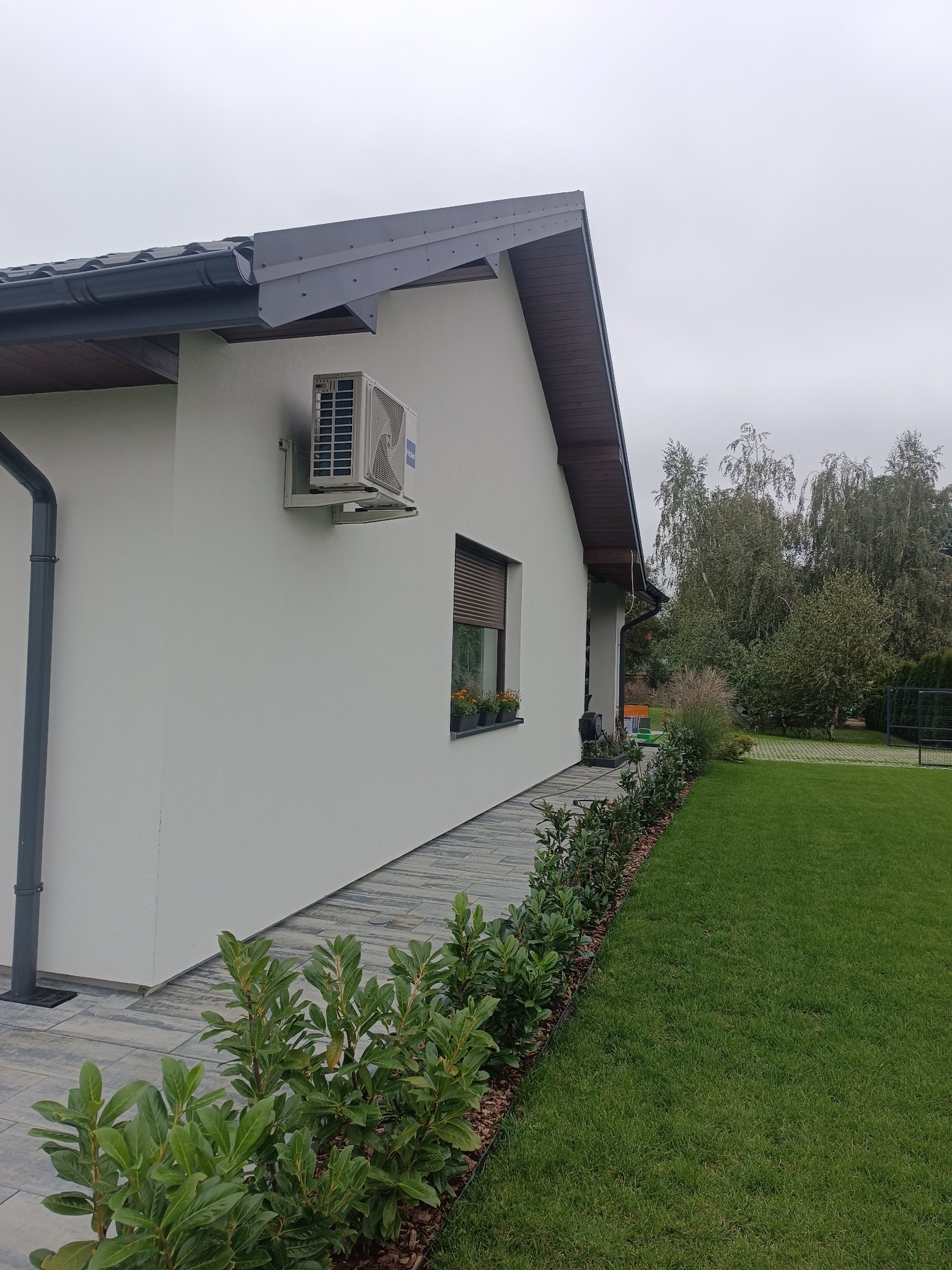 Boczny widok domu z jasną elewacją, klimatyzatorem na ścianie i oknem z żaluzjami. Wzdłuż ściany biegnie ścieżka z kostki, a obok niej zadbany trawnik i krzewy.